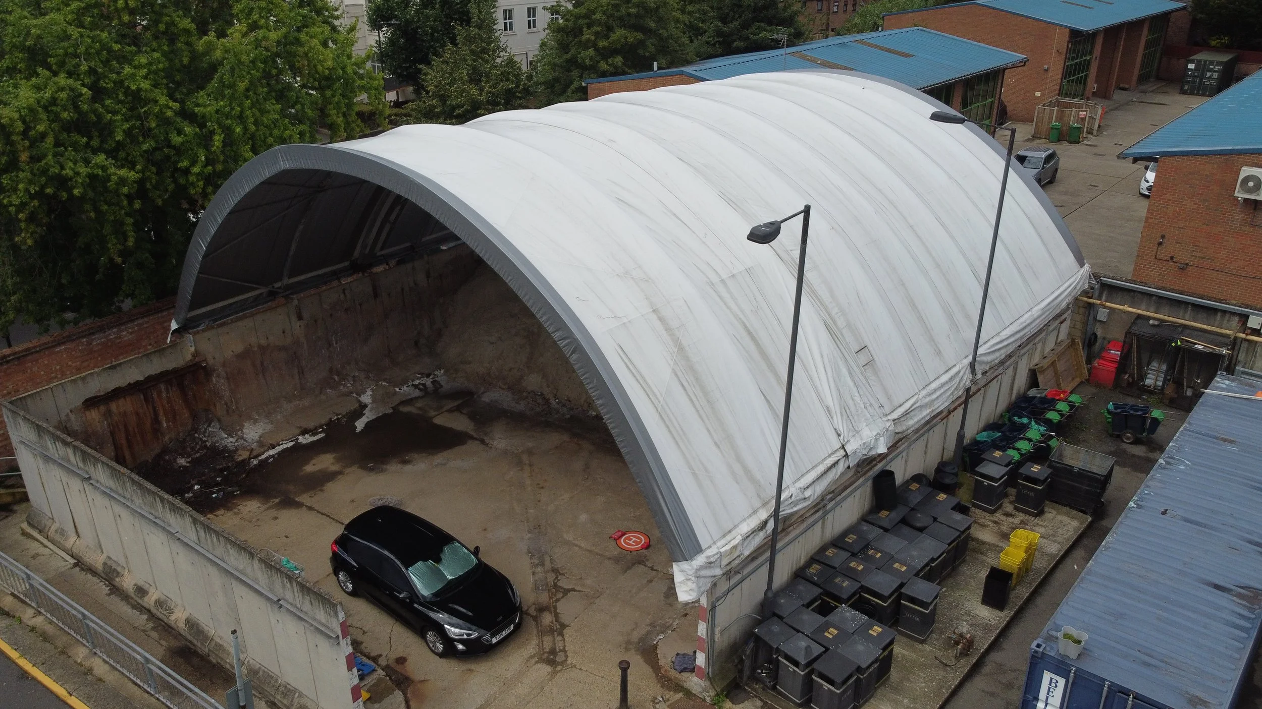 An aerial view of a large covered parking area with a curved roof, parked cars, green trees, and surrounding buildings with blue roofs.