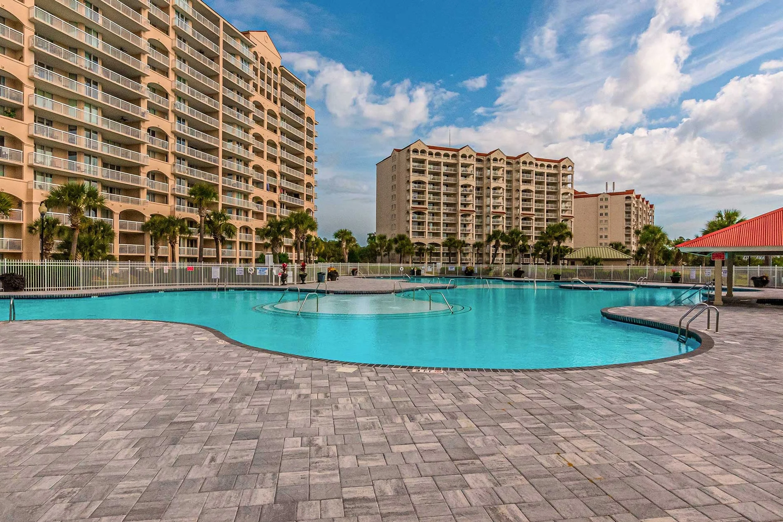 A view of several high-rise residential buildings with balconies, a swimming pool in the foreground, and palm trees around the pool area. The sky is partly cloudy.