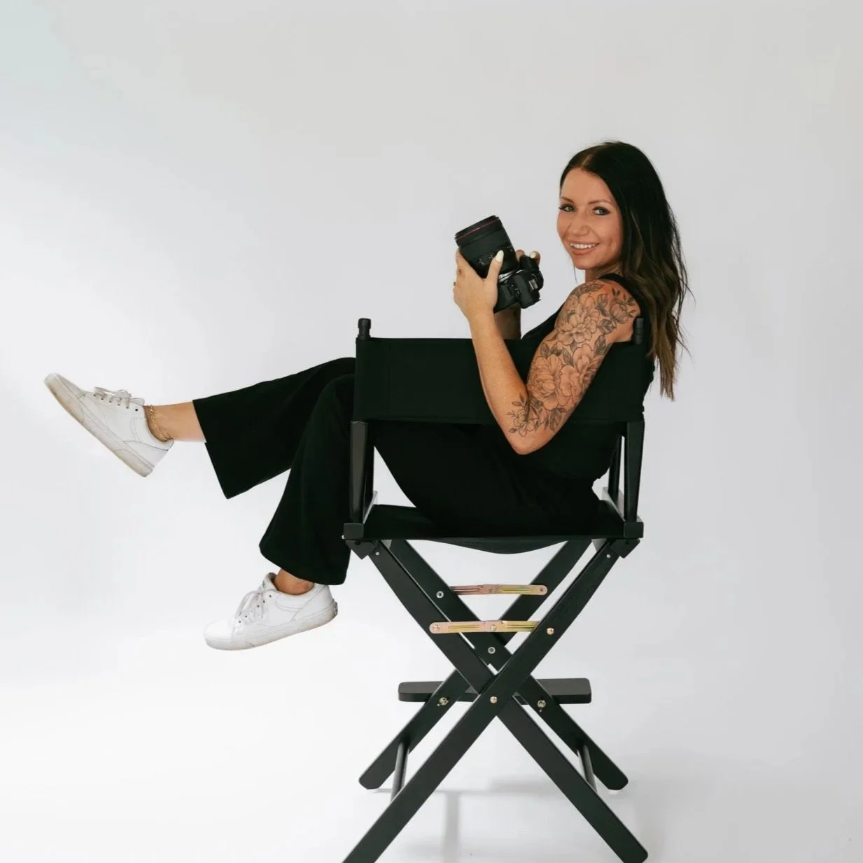 A woman with dark hair in a black outfit sitting on a stool, holding a camera, smiling against a plain background.