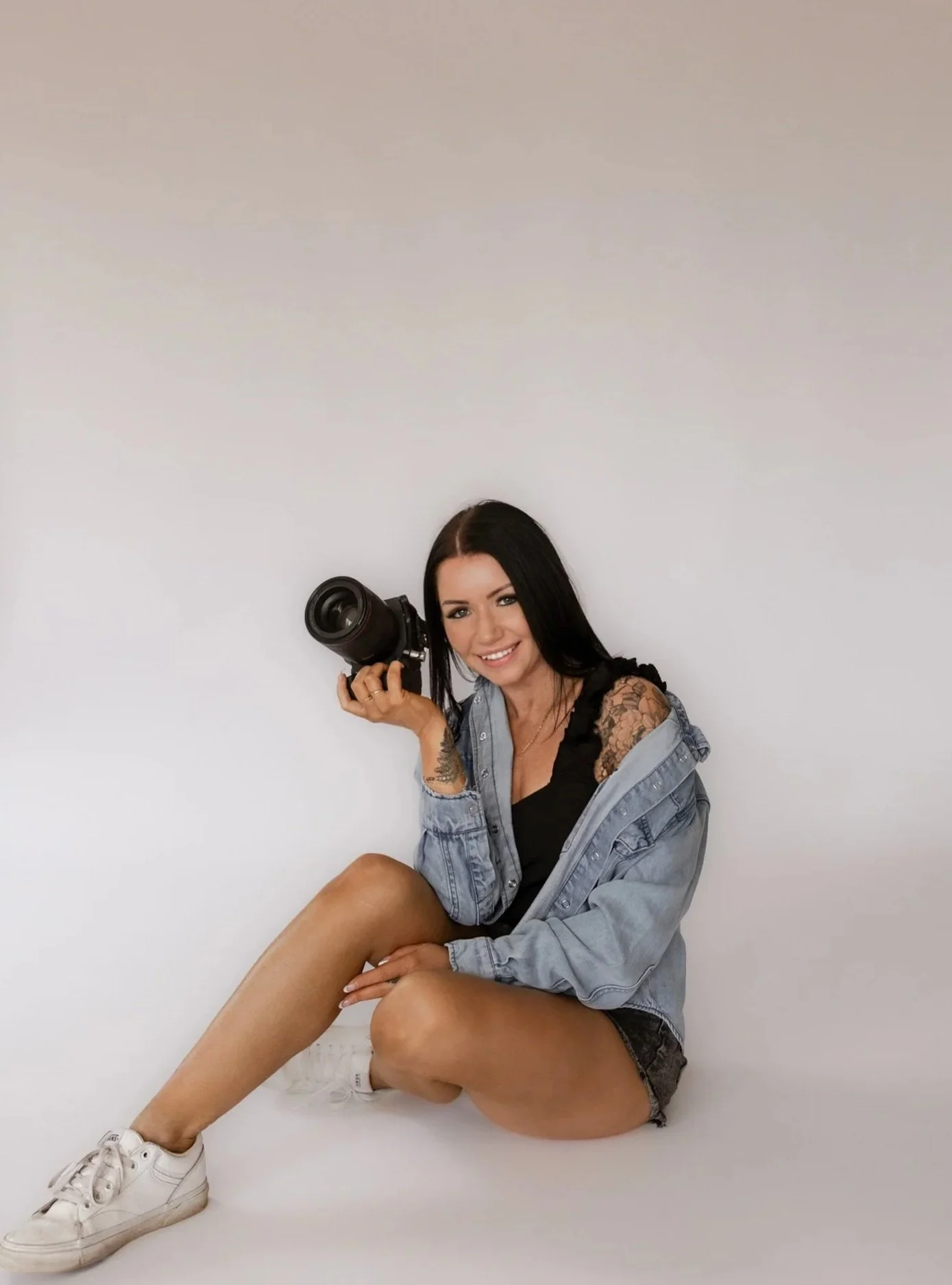 A woman with dark hair in a black outfit sitting on a stool, holding a camera, smiling against a plain background.