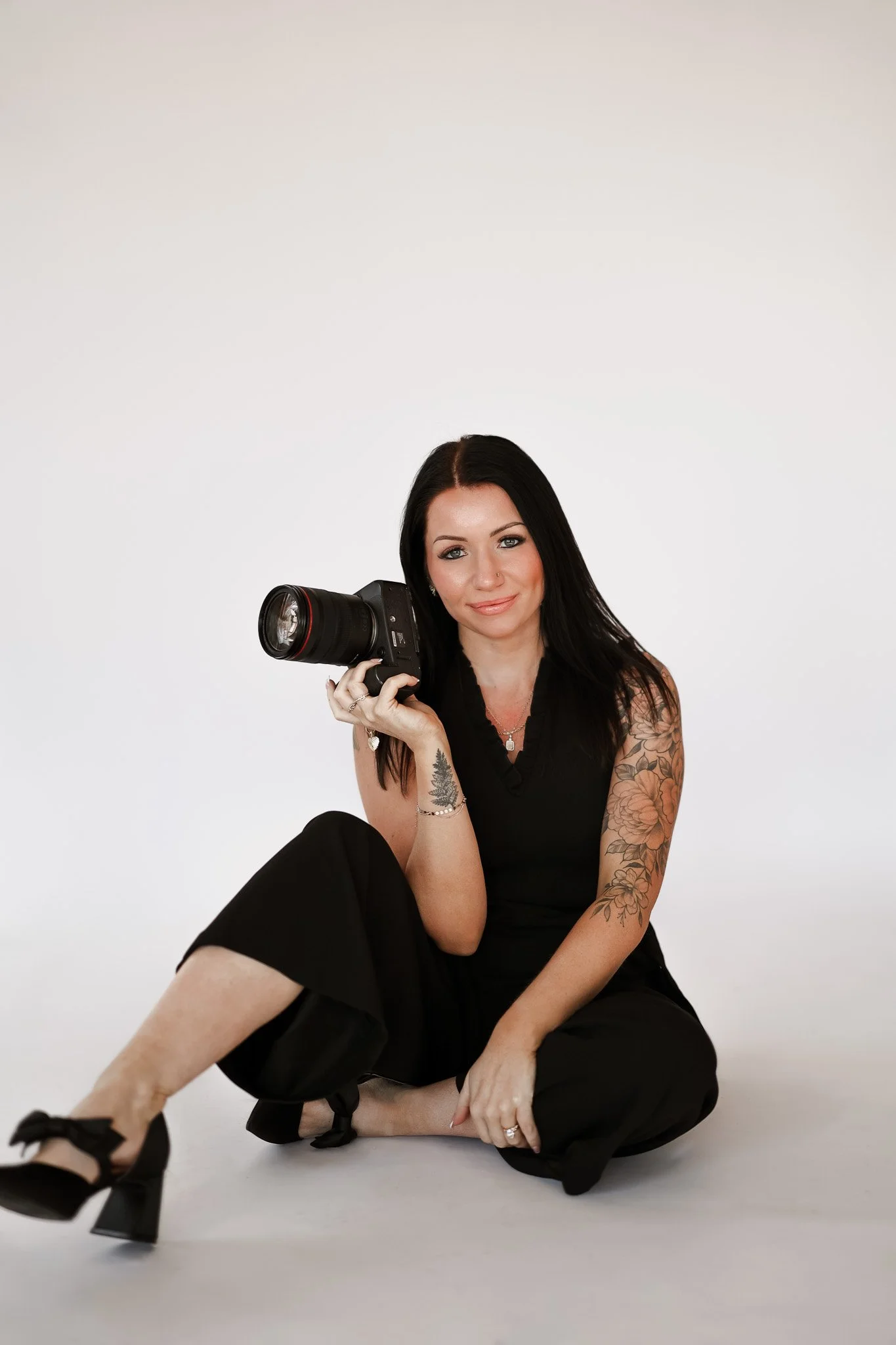 A woman with dark hair in a black outfit sitting on a stool, holding a camera, smiling against a plain background.