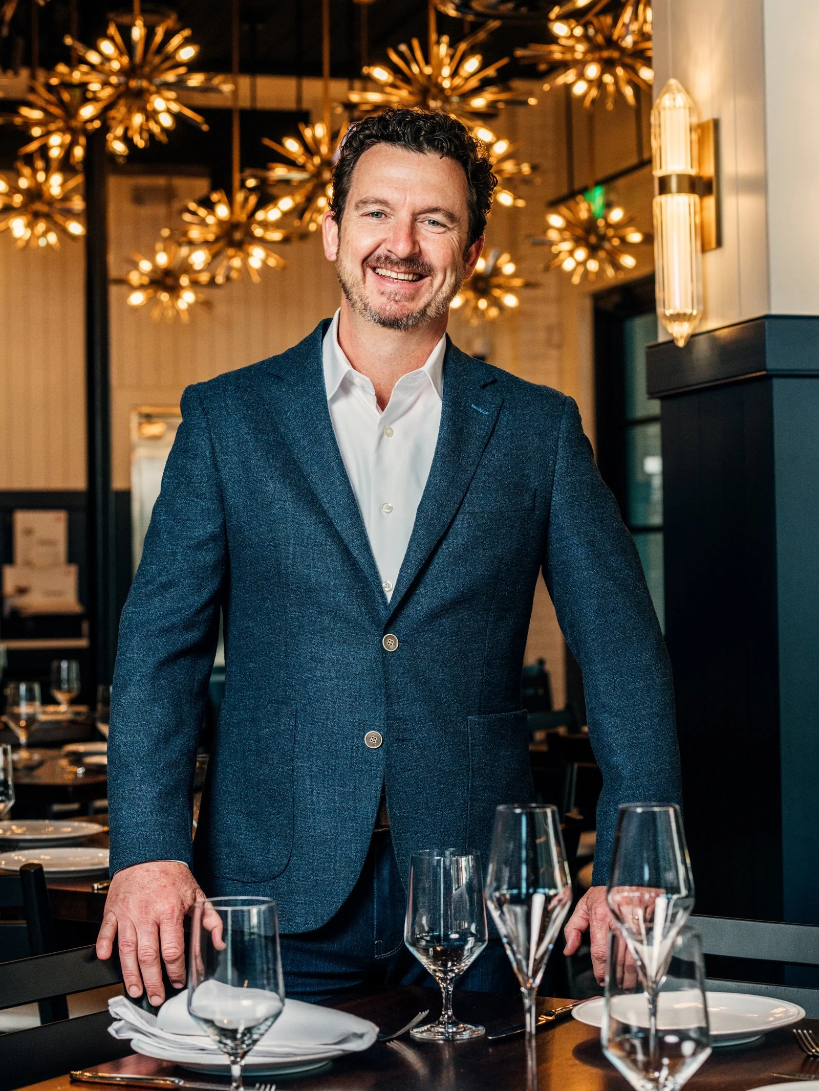 A man in a blue blazer and white shirt standing at a restaurant table, smiling with his hand resting on the table, in a stylish restaurant with modern lighting fixtures.