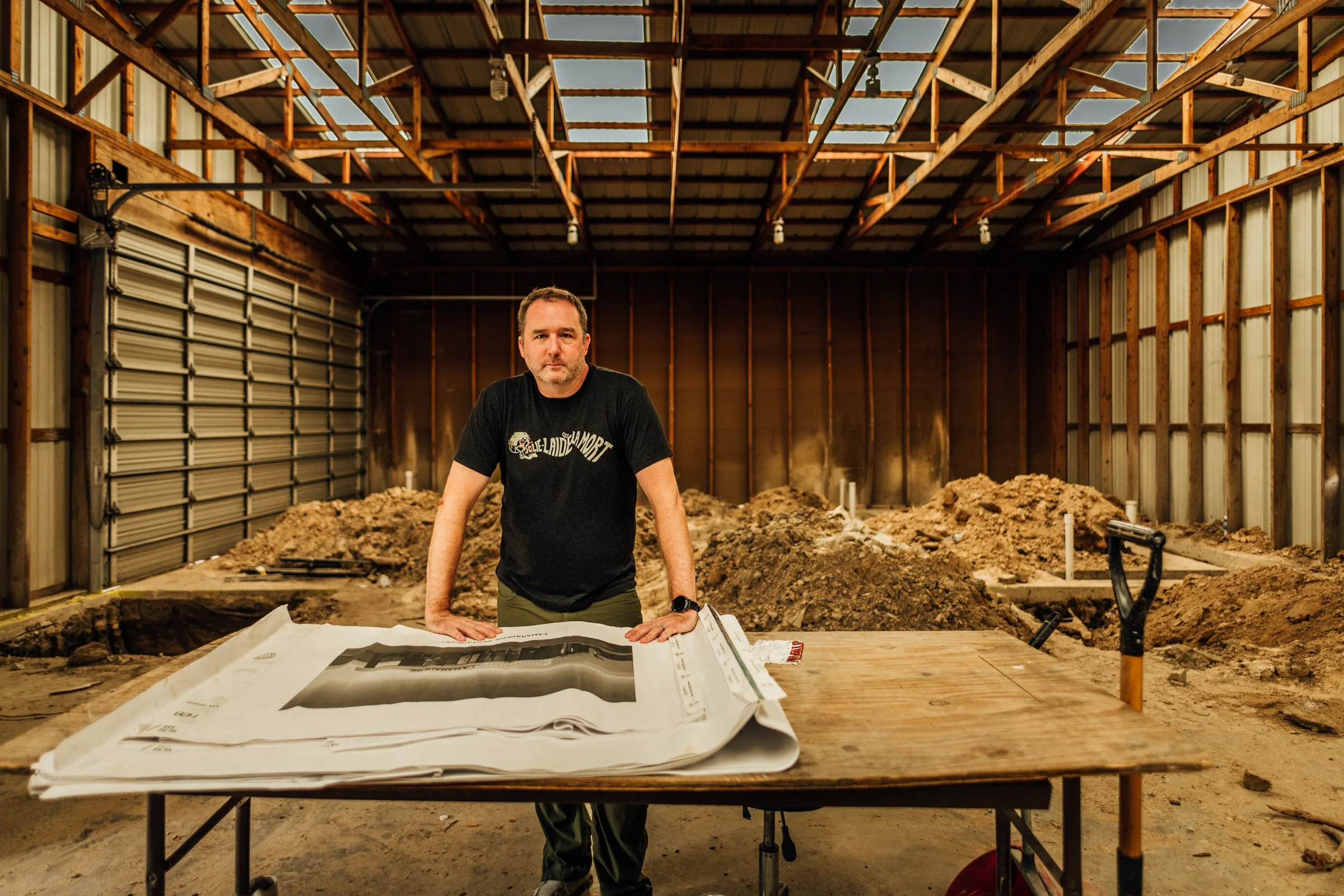 Man standing inside a partially constructed building with dirt piles, tools, and blueprint on a table.