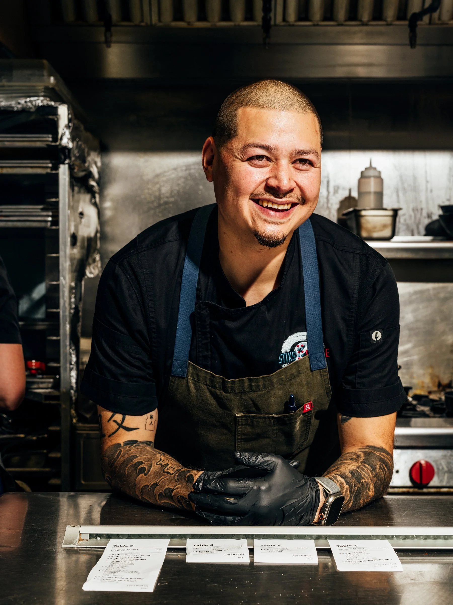 A smiling male chef with tattoos on his arms, wearing a black shirt, an apron, and black gloves, standing behind a counter with restaurant tickets in front of him in a commercial kitchen.