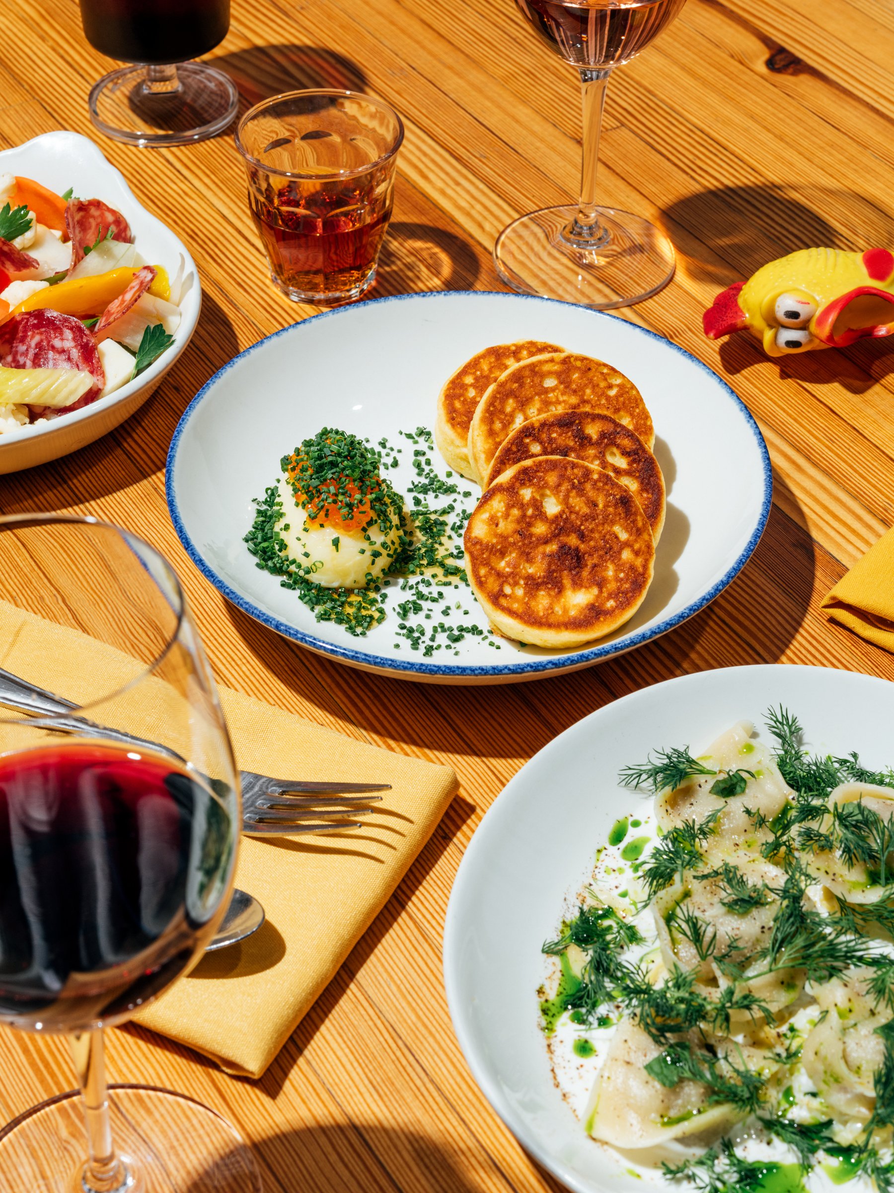 A table set with various dishes including a plate of four small pancakes with mashed potatoes and chives, a bowl of potato salad with vegetables, a plate of ravioli with herbs, glasses of red and white wine, and a glass of rosé, with a wooden table a