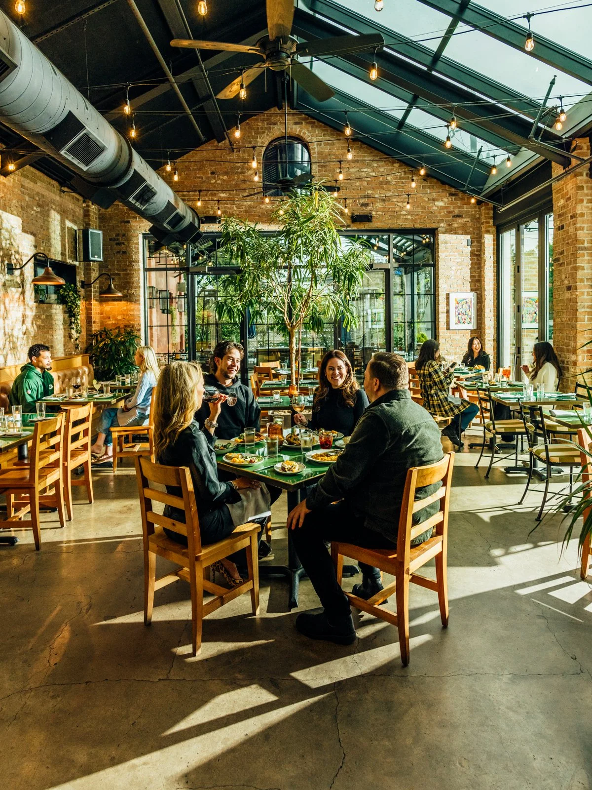 Group of five people enjoying a meal together at a restaurant with large windows, exposed brick walls, and a high glass ceiling with string lights.