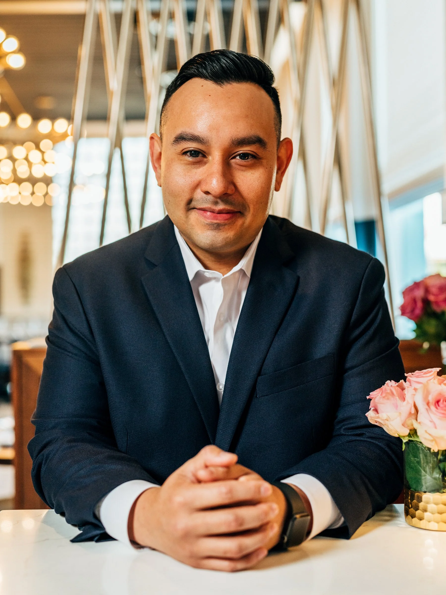 Man in a dark suit with white shirt sitting at a table with folded hands, smiling at the camera, in a modern restaurant or cafe with flowers on the table.