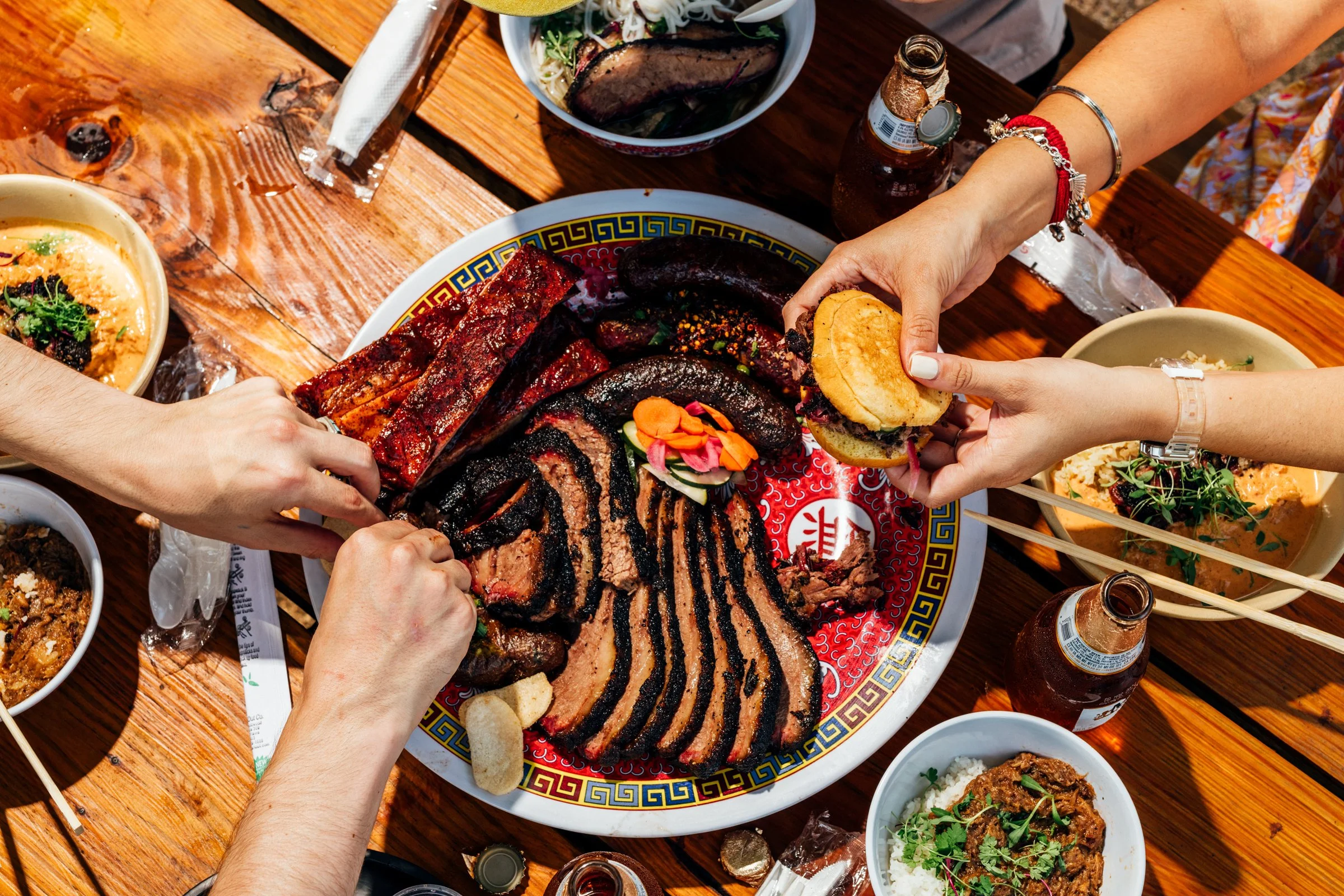 People sharing a platter of assorted grilled meats, including ribs and sausage, with sides of rice and salads on a wooden table, with drinks and condiments visible.