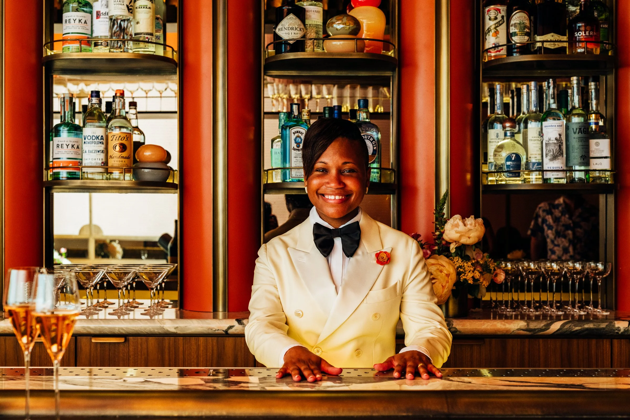Smiling female bartender with short hair, dressed in a white tuxedo jacket with a black bow tie, standing behind a bar with bottles of alcohol on shelves in the background.