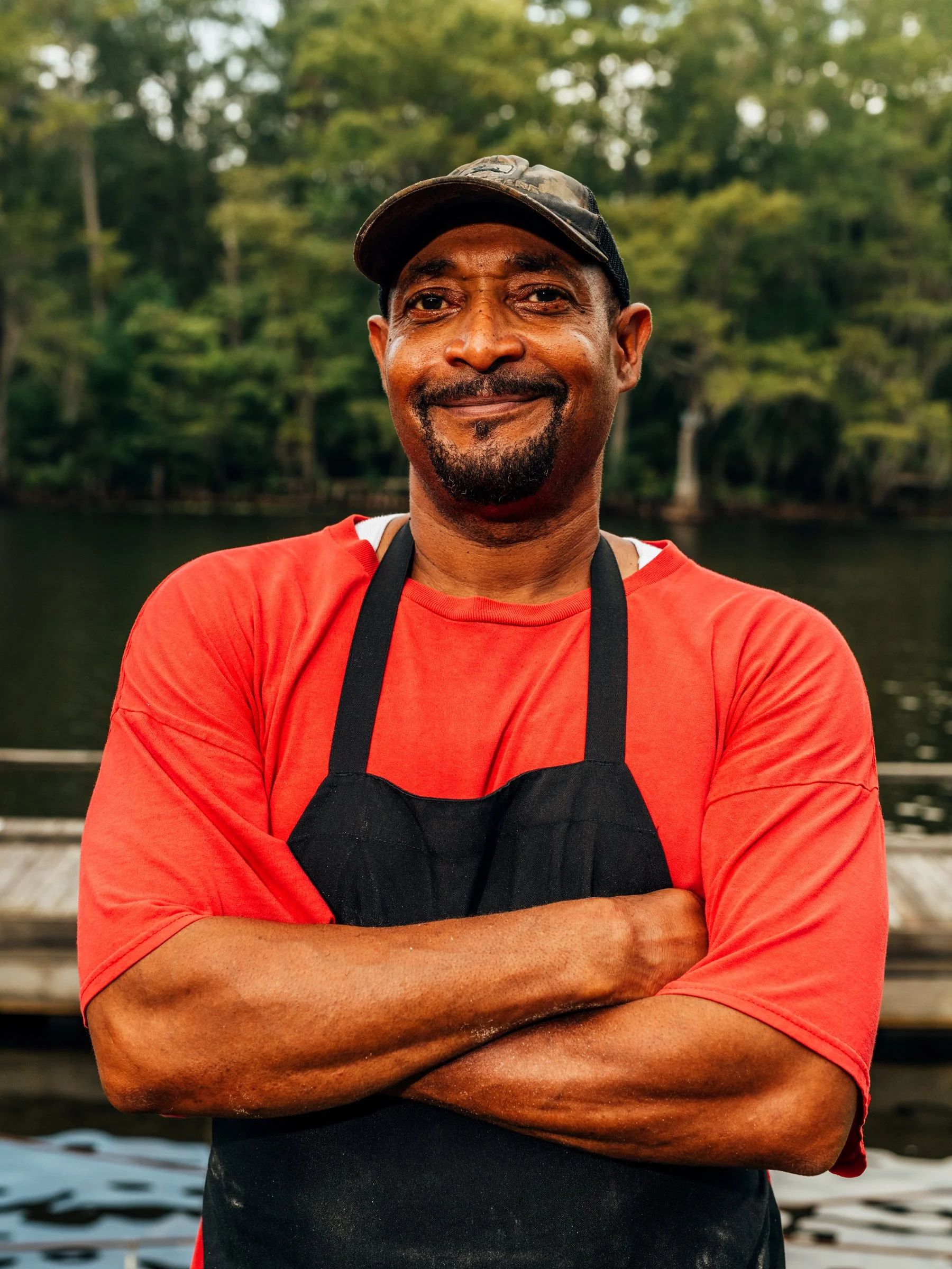 A man in a red shirt and black apron standing outdoors near water, smiling with arms crossed, with green trees in the background.