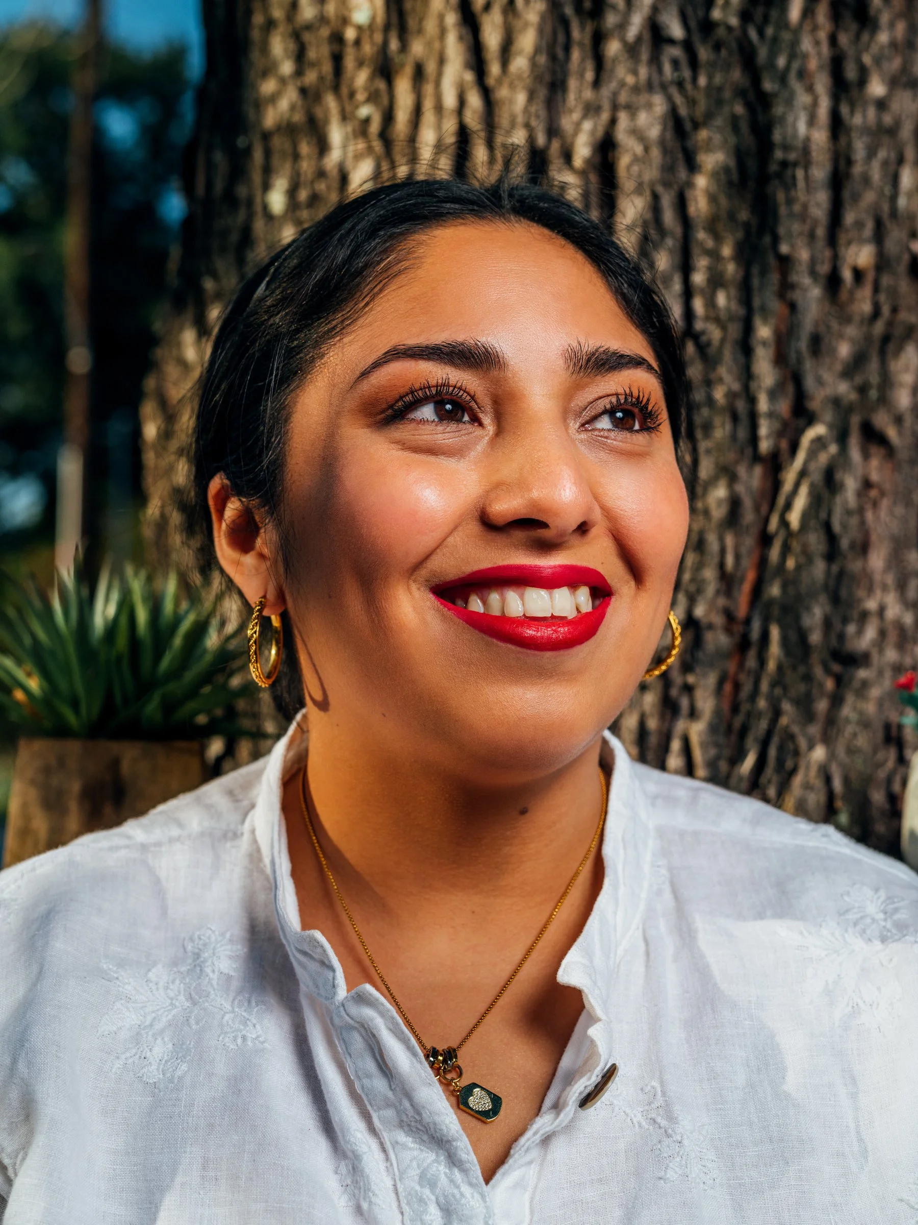A smiling woman with dark hair, red lipstick, wearing gold hoop earrings, a gold necklace with a pendant, and a white shirt, standing outdoors near a large tree.