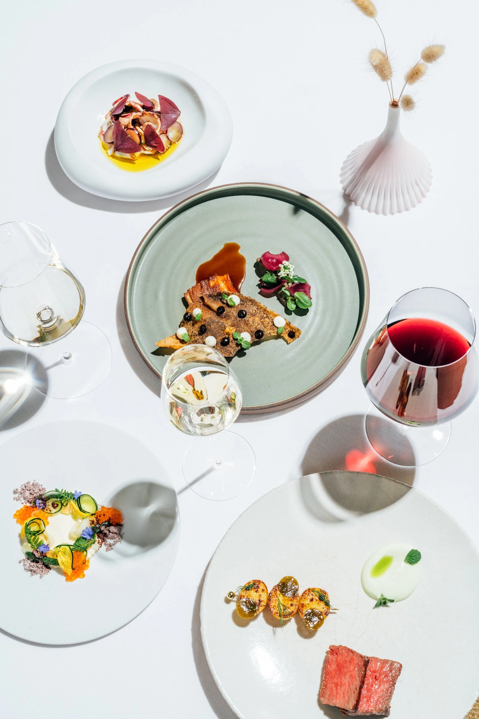 Assorted gourmet dishes and glasses of wine on a white table, including a plate with a slice of meat with sauce and garnishes, a small plate with colorful vegetables, and a white ceramic plate with multiple small food items, plus a white vase with dr