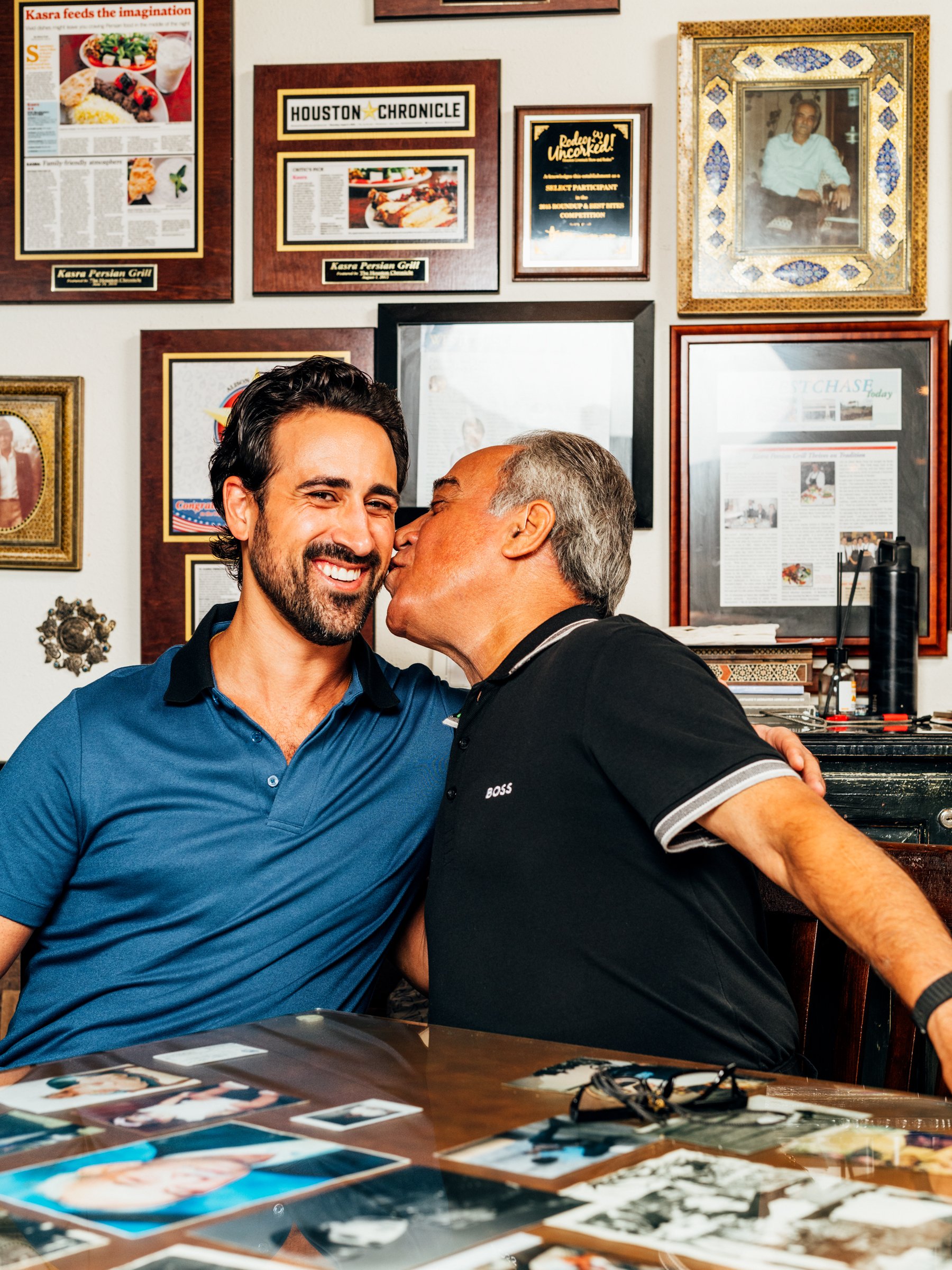 Two men sharing an affectionate moment, one kissing the other's cheek, in a room decorated with framed photos, magazine articles, and awards.