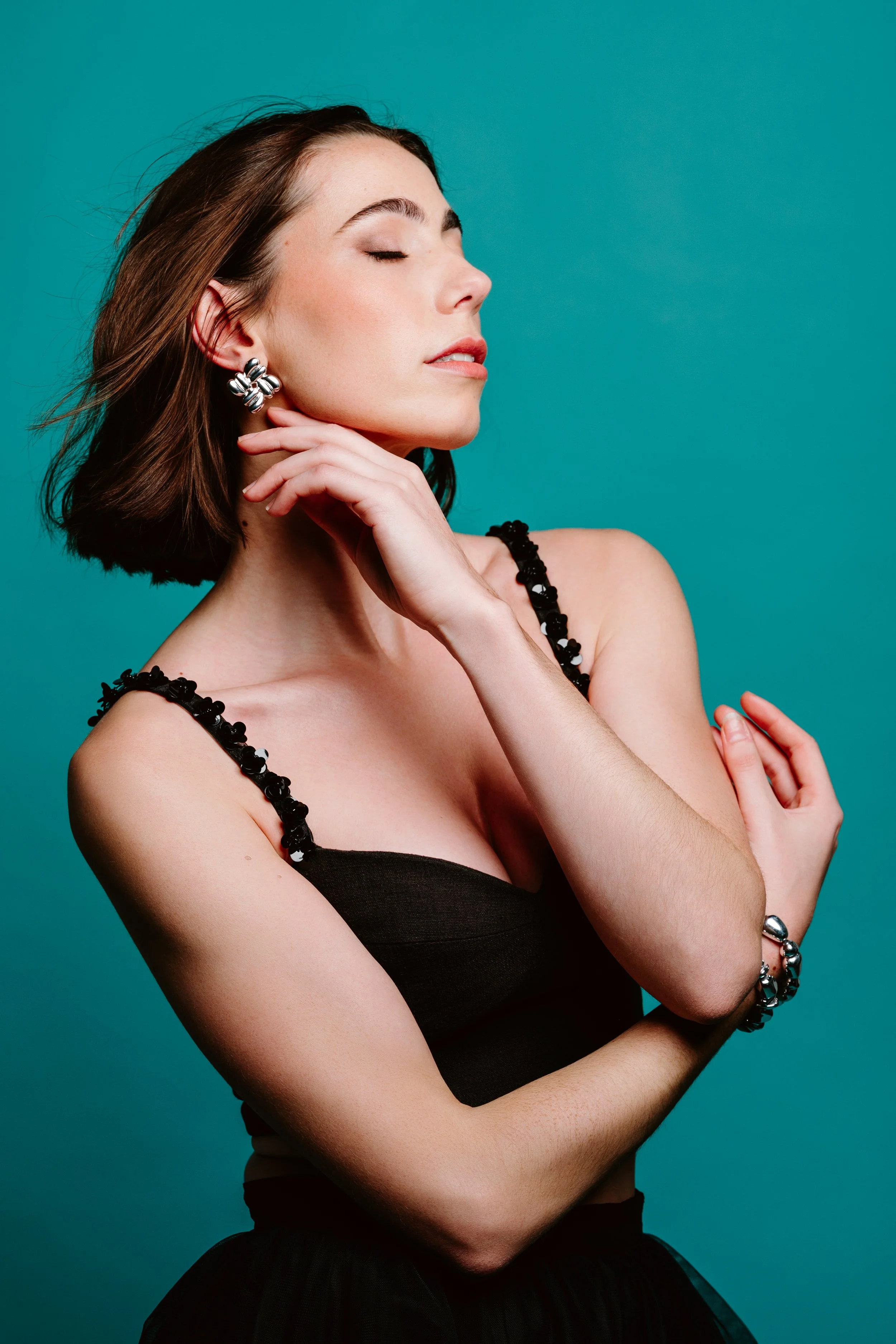 A woman with brown hair wearing makeup and gold earrings, dressed in a black textured top, looking down against a pink background.