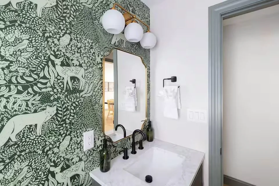 A bathroom with blue and white patterned wallpaper, white towels hanging on black hooks, a small wooden bench with rolled towels, a plant in a pot, a framed nature photograph, and a partial view of a white vanity with a sink.