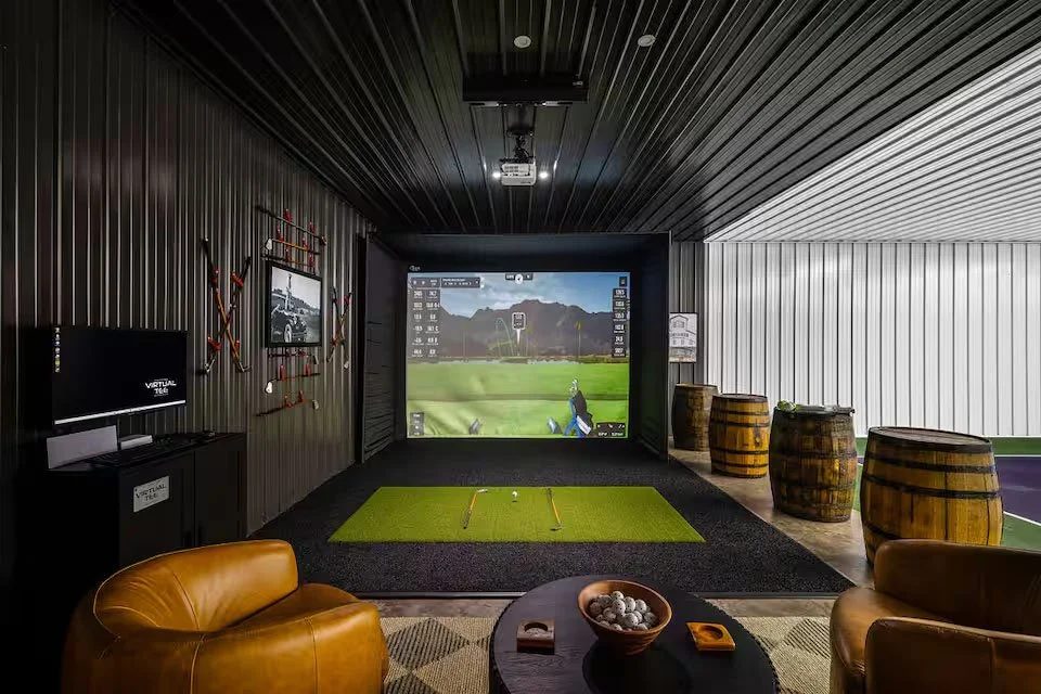 A cozy game room with a pool table, dartboard, and Scottish checkers on the wall. There is a Tiffany-style lamp hanging from the wooden ceiling, and a large window with white curtains allowing natural light in. The room has wood-paneled walls and hardwood floors, with a small table and chairs in the corner.