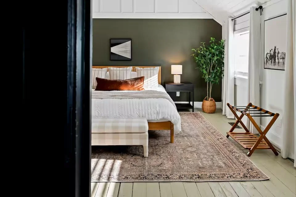 A bedroom with a dark wooden bed frame, beige and green bedding, and four pillows. There are two nightstands with lamps on each side of the bed. The room has a ceiling fan with wooden blades. Green curtains cover two windows, and a framed artwork hangs on the wall. There is a gray armchair with a white pillow near a large potted plant. The floor is hardwood, and an area rug covers part of it.