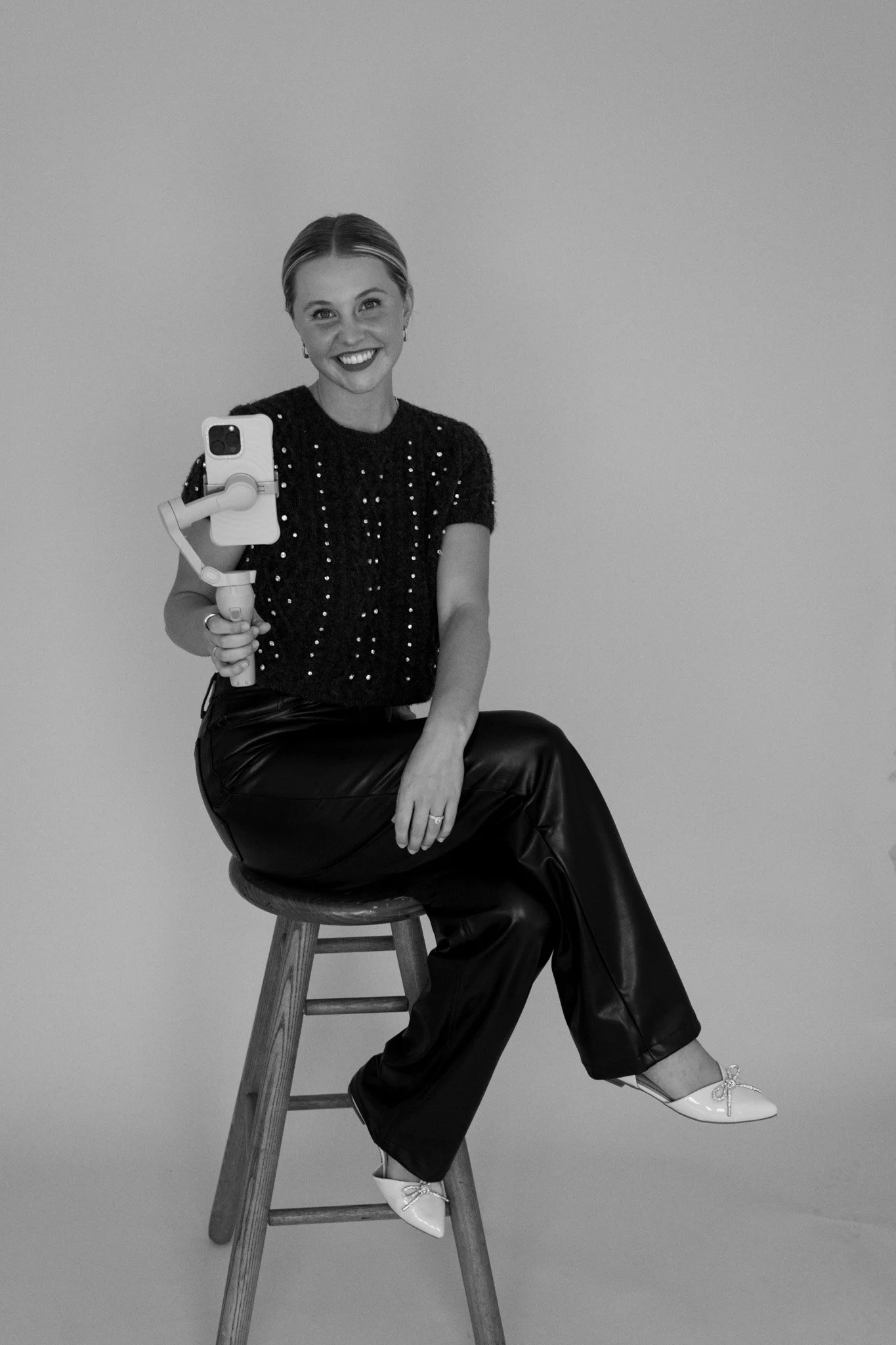 A woman sitting on a stool, smiling, holding a phone mounted on a stabilizer, wearing a short-sleeved, beaded top, leather pants, and white shoes, against a plain background.