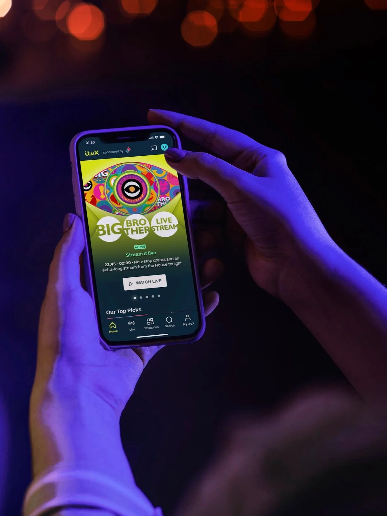 Hands holding mobile device showing the Big Brother Live Stream hero on the ITV homepage including a CTA to 'Watch Live'