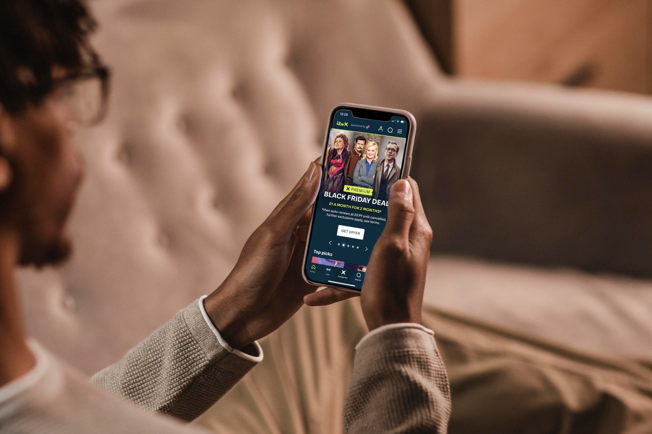 Man holding a mobile displaying the Black Friday offer homepage hero featuring the selected promo talent: Nolly, Parks & Recreation and A Spy Amongst Friends