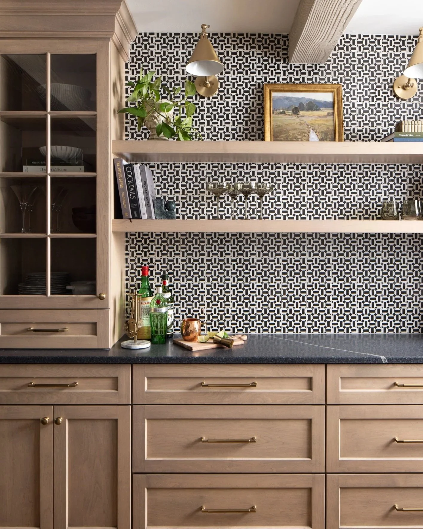 A kitchen with light wooden cabinets and a black countertop. Two floating shelves hold books, drinking glasses, and decorative items. A backsplash has a black and white geometric pattern. Two brass wall sconces, a painting, and a potted plant are on the wall.