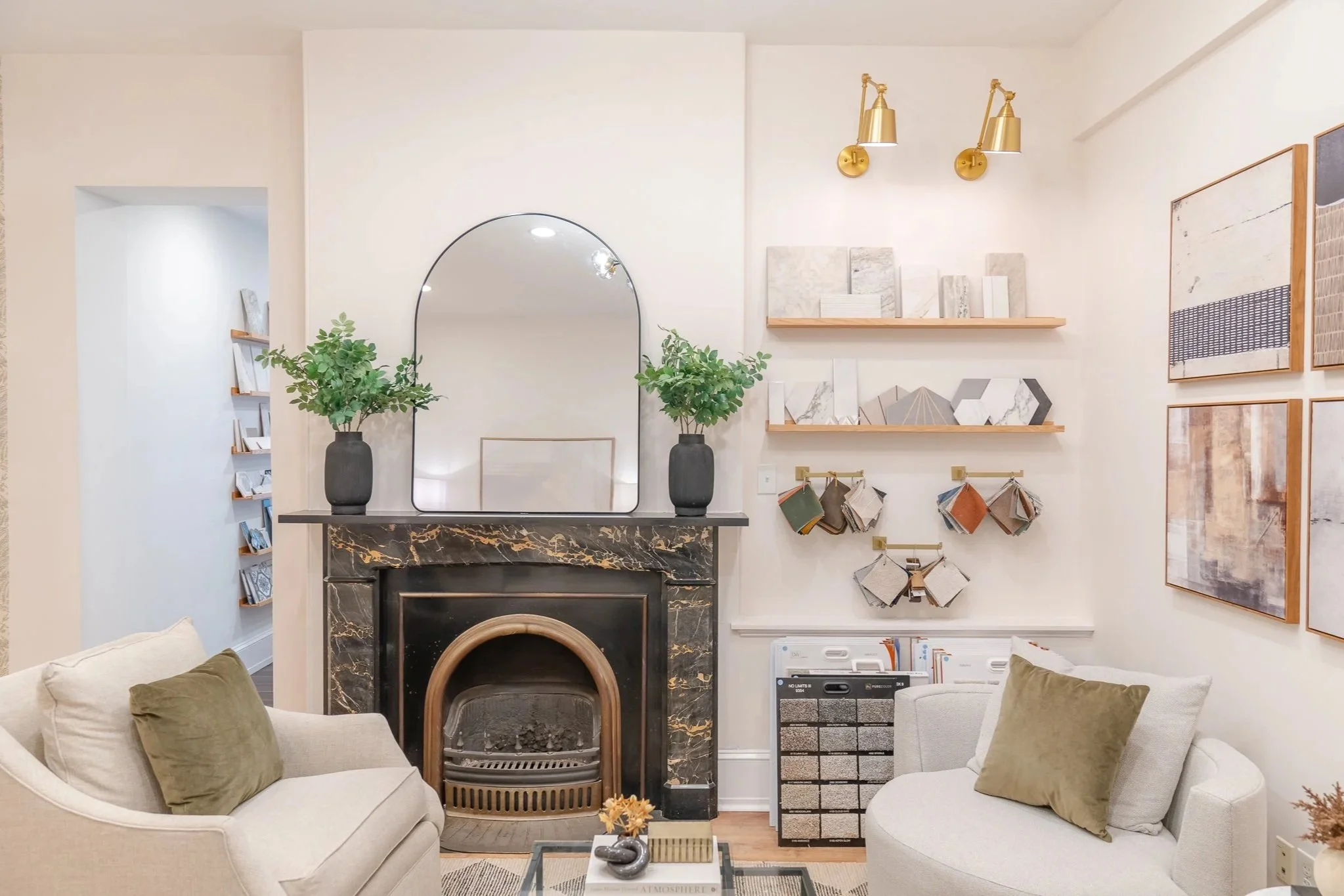 Living room with a black marble fireplace, gold wall lamps, decorative artwork, and a mirror above the fireplace. Two beige sofas with green pillows, a coffee table with decor items, and shelves with fabric samples and artwork.