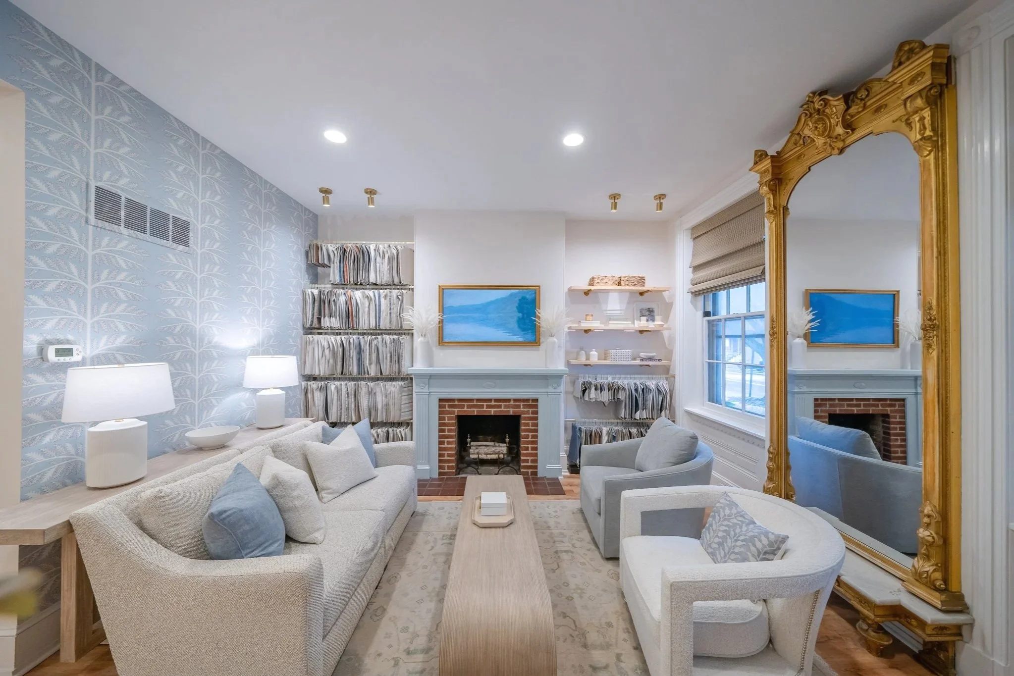 Living room with a beige sofa and armchair, a brick fireplace, a large gold mirror, two lamps, a flatscreen TV, bookshelves with decorative items, and windows with beige shades.