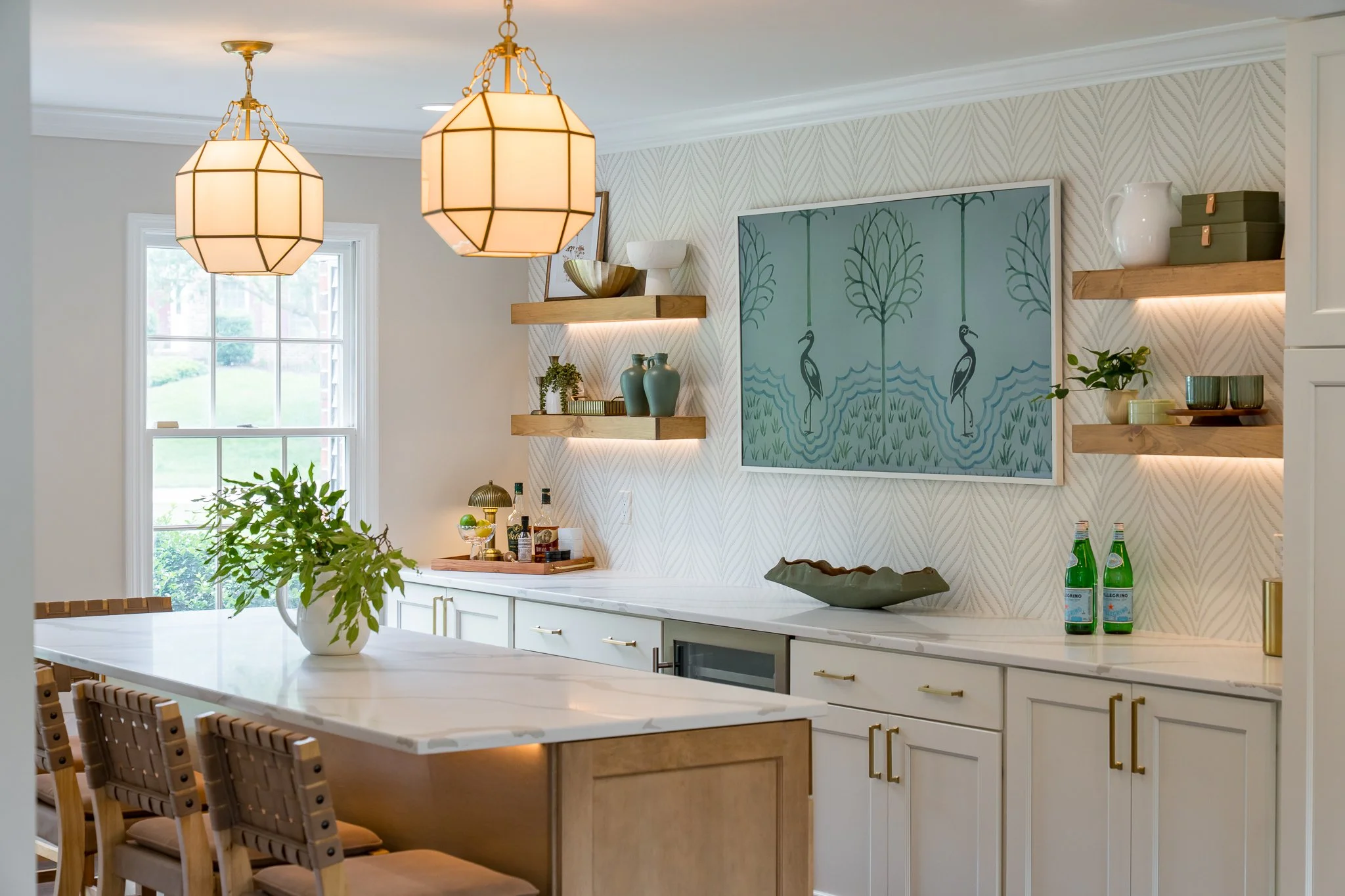 Bright kitchen with white cabinetry, marble countertops, and wooden open shelves displaying decorative items and plants. Two pendant lights hang above a large island with wooden chairs. A window provides natural light, and a blue artwork with crane images hangs on the patterned wall.