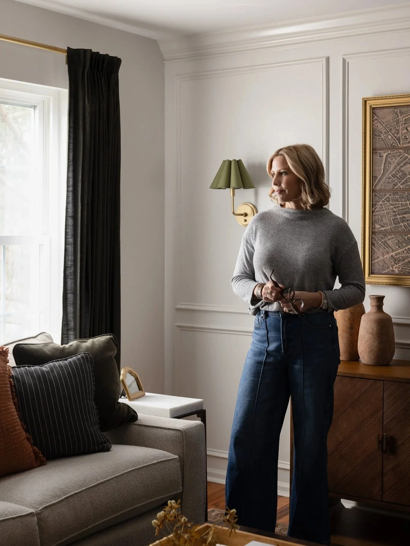 A woman with shoulder-length blonde hair, dressed in a gray sweater and blue jeans, standing in a well-lit living room holding glasses, gazing out of a window with dark curtains.