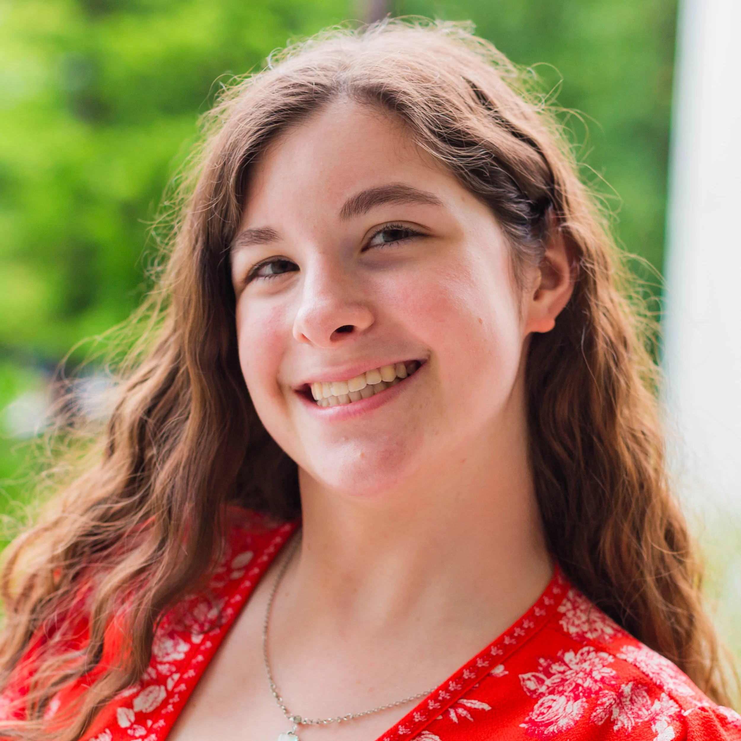 A caucasian young adult female with long wavy brown hair smiles are the camera. She is wearing a red floral top