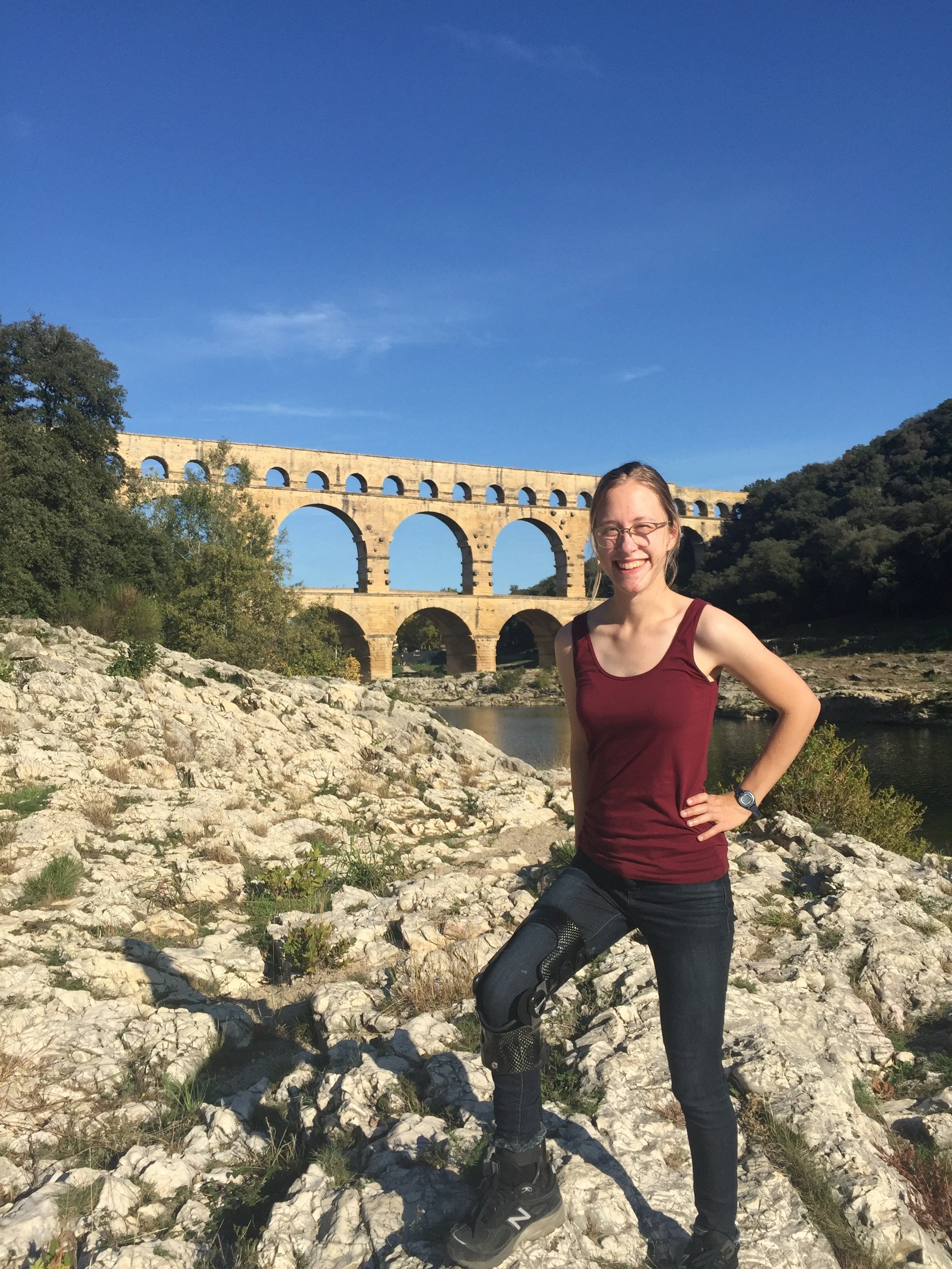 a white woman standing on rocky terrain outdoors near a body of water with a bridge in the background