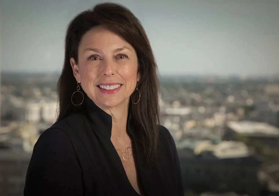 A woman with dark brown hair, wearing a black top and hoop earrings, smiling in front of a cityscape background.