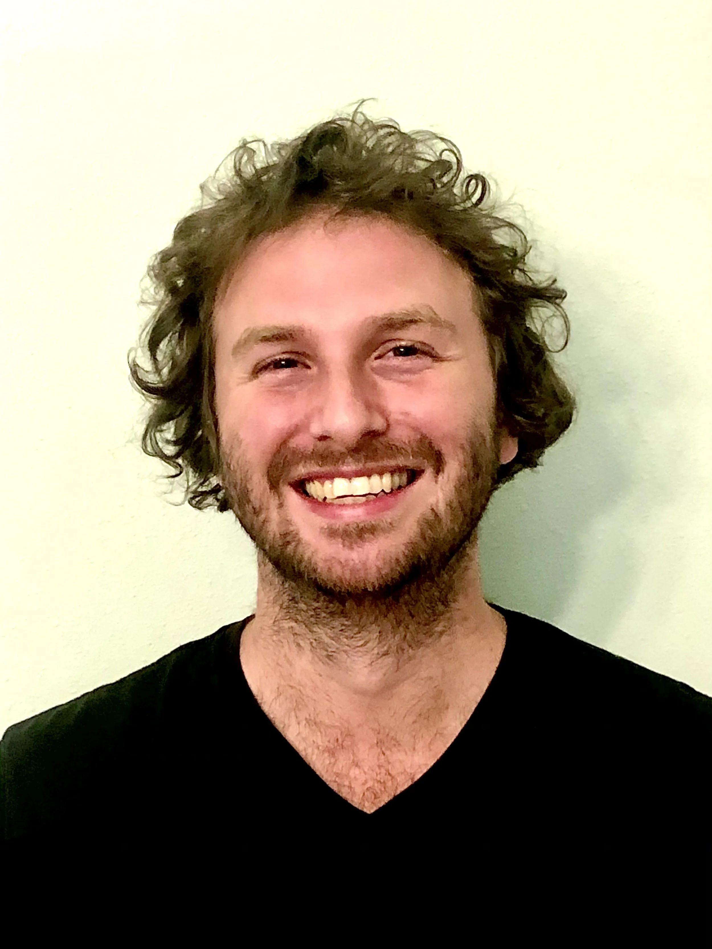 A smiling man with curly brown hair and a beard, wearing a black shirt, standing against a light-colored wall.