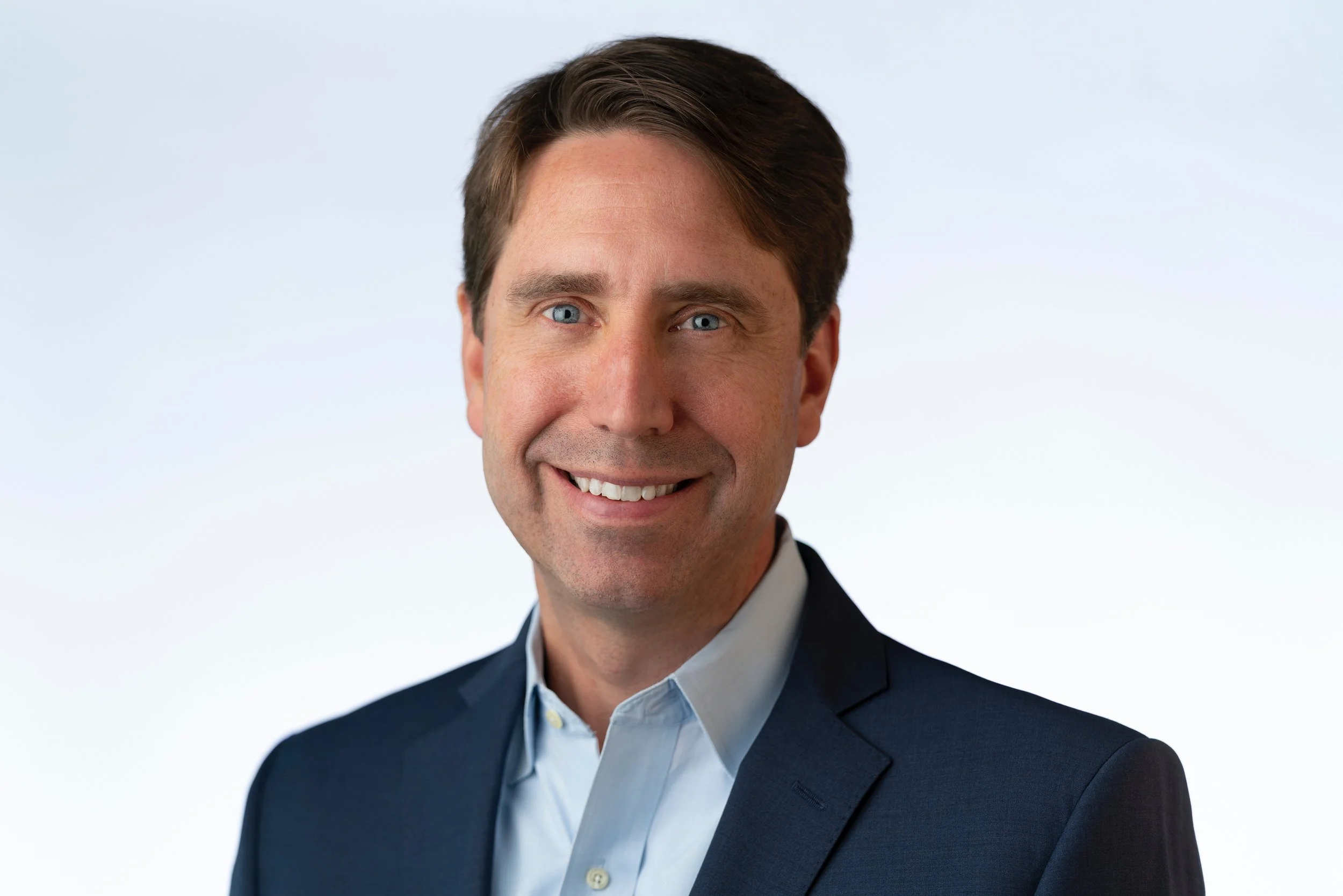 A man with brown hair and blue eyes wearing a dark suit and light dress shirt, smiling at the camera.