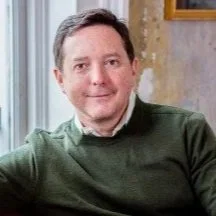 A man with short brown hair and a light complexion, wearing a green sweater, sitting indoors near a window with a curtain.