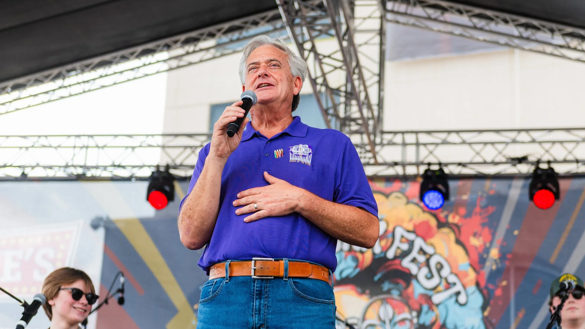 An older man with gray hair speaking into a microphone on a stage at an outdoor event, with a woman and another person visible in the background, and a colorful background banner.