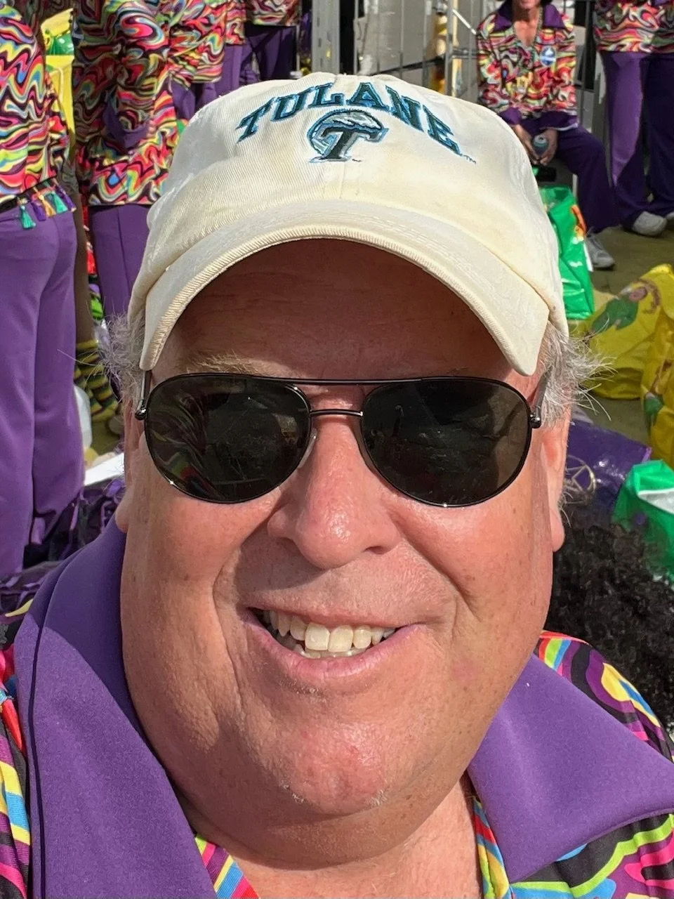 Close-up of a smiling man wearing sunglasses, a white cap with 'TULANE' embroidered on it, and a colorful patterned shirt with purple accents. People in similar colorful outfits are in the background at an outdoor event.