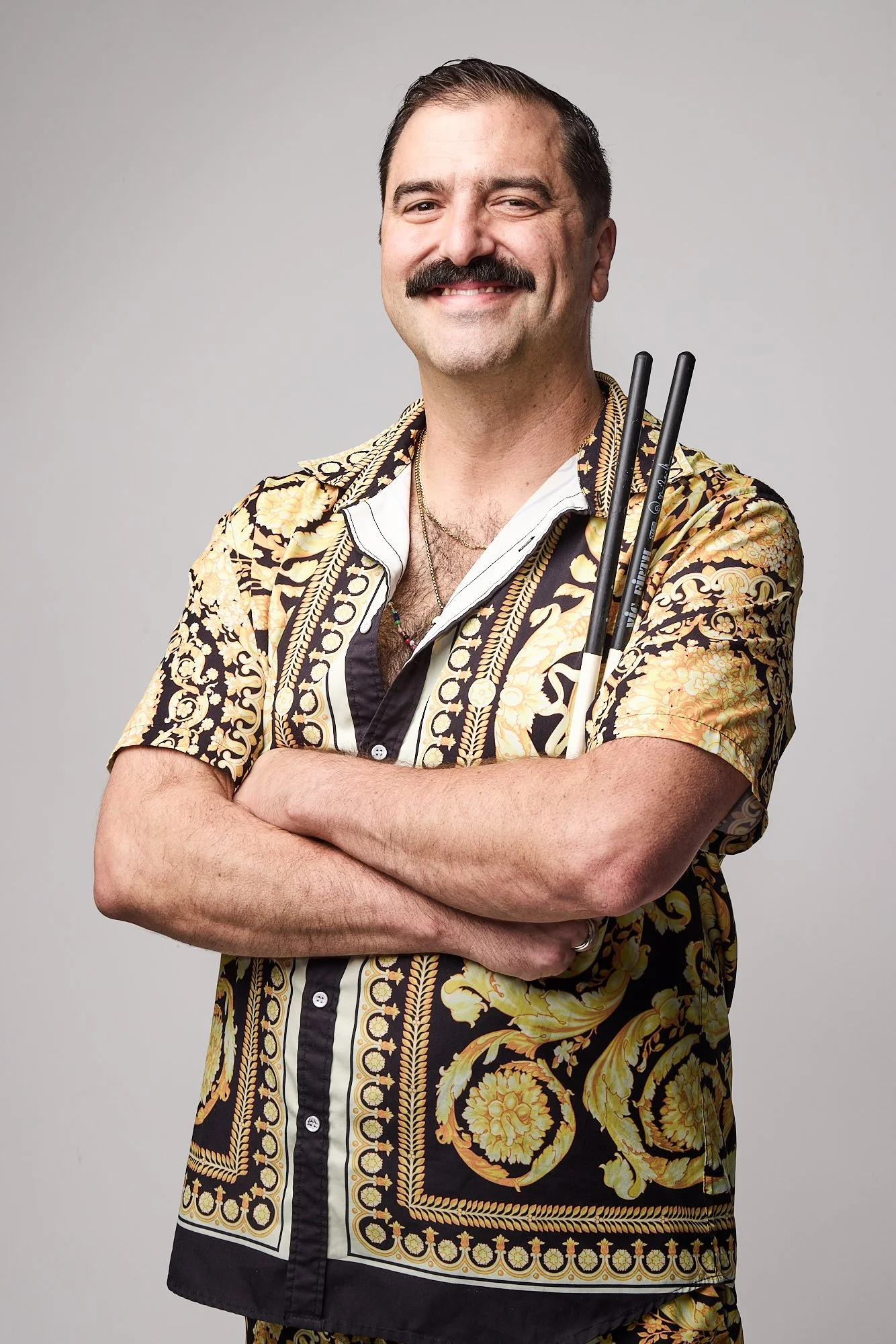 A man with a mustache, wearing a patterned shirt, arms crossed, smiling, holding drumsticks.