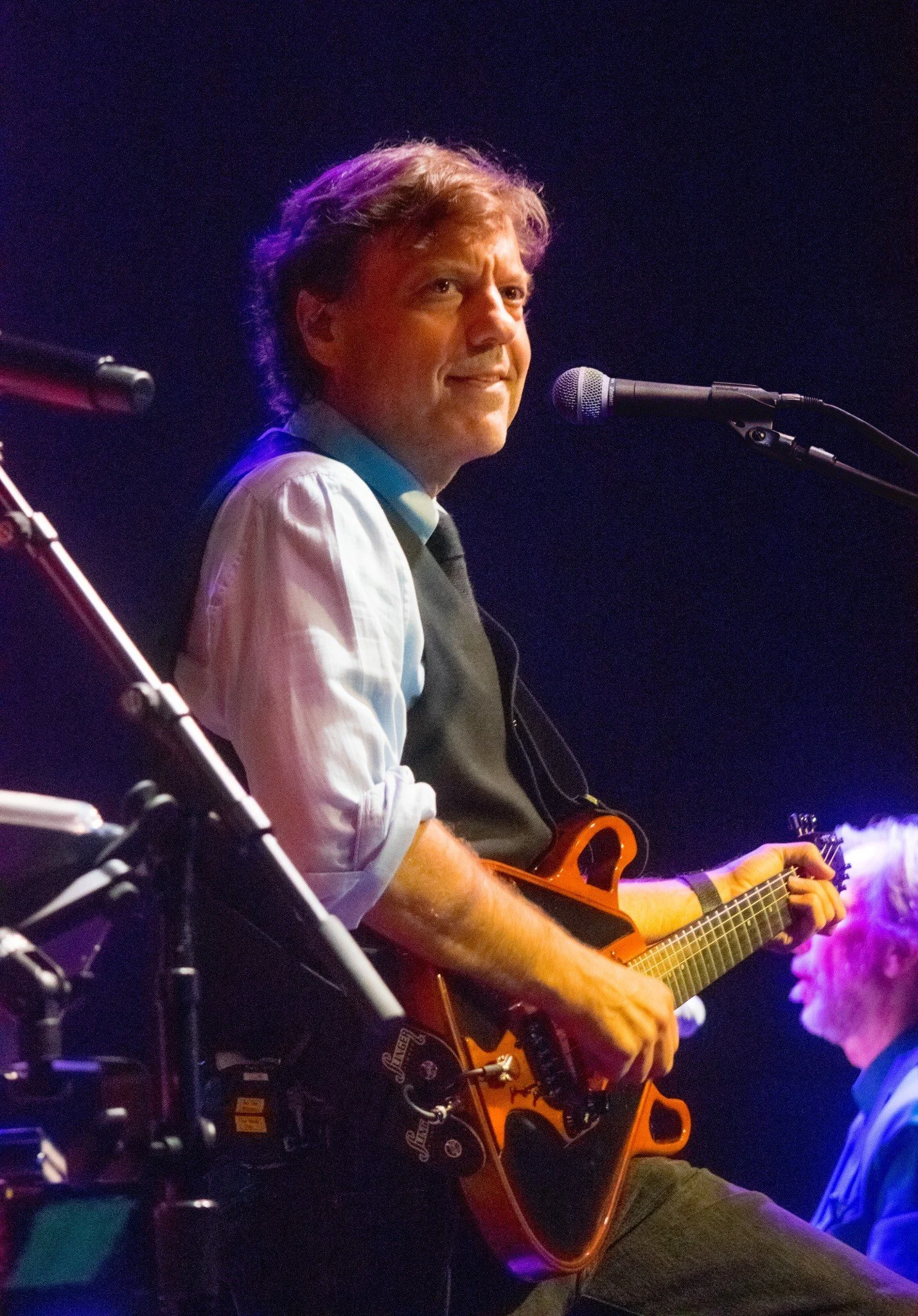 A man playing an electric guitar on stage, seated near a microphone with a dark background.