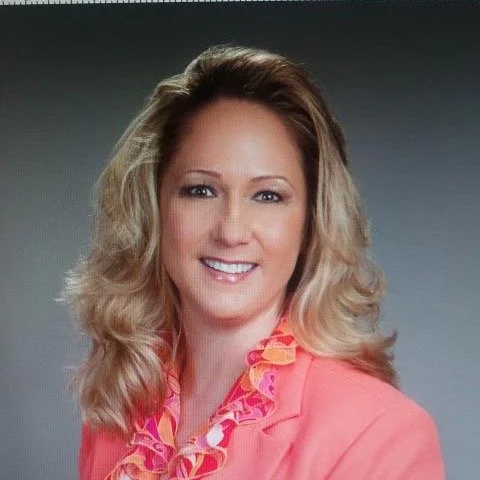 A woman with blonde wavy hair, wearing a coral blazer and a pink patterned blouse, smiling at the camera against a gray background.