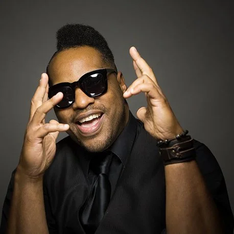 A man wearing black sunglasses, a black shirt, and a black tie, smiling and gesturing with both hands near his face.