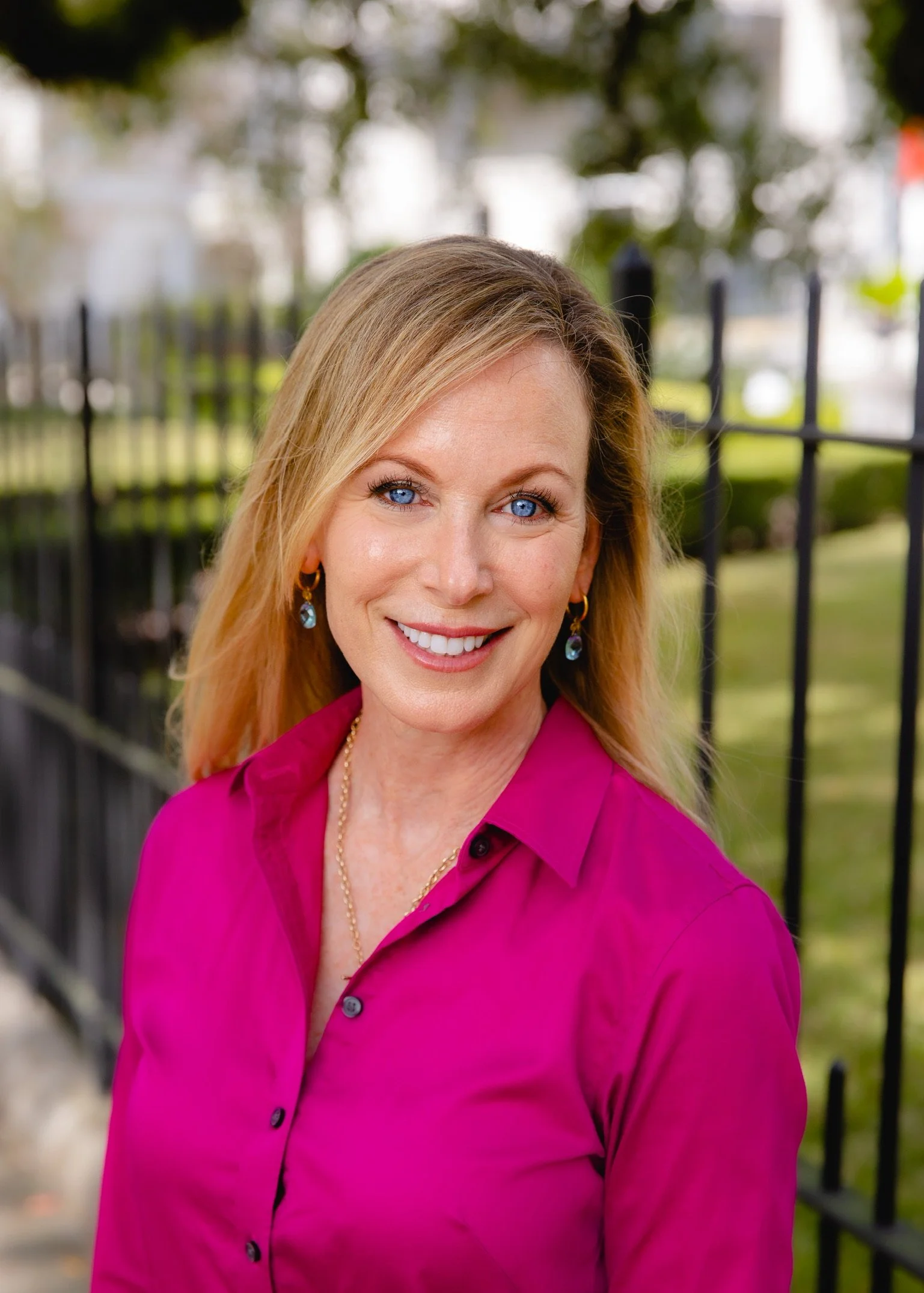 A smiling woman with blue eyes and blonde hair, wearing a pink blouse, earrings, and a gold necklace, outdoors with a black fence and trees in the background.