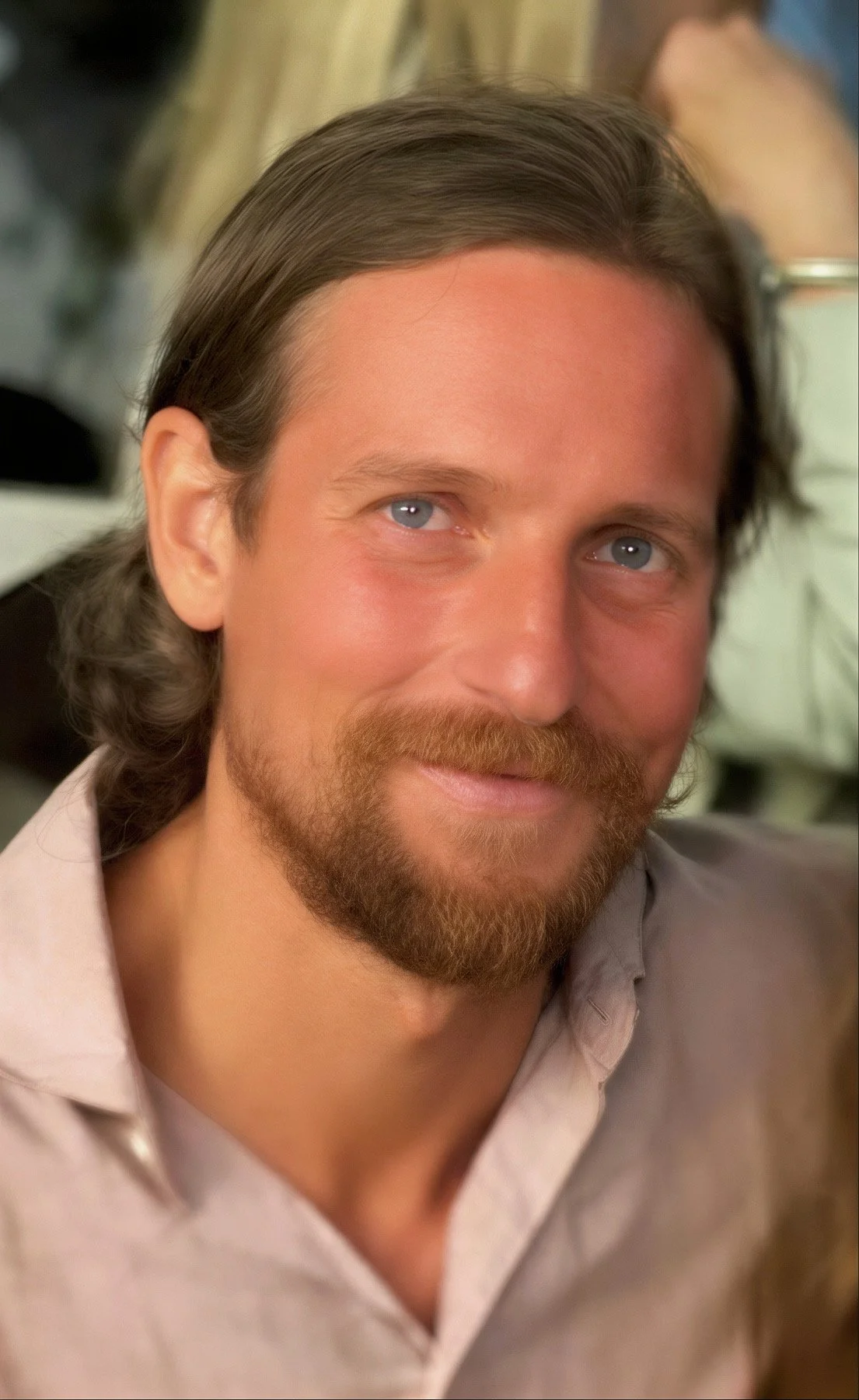 Close-up of a man with long brown hair, blue eyes, and a full beard, smiling softly, wearing a light beige shirt.