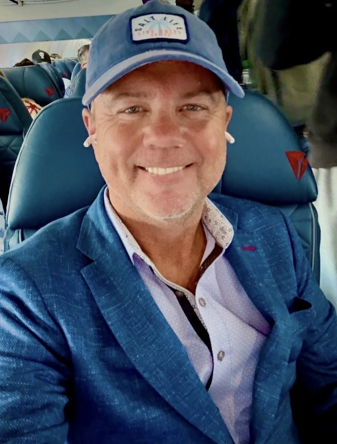 Smiling man in a blue jacket and baseball cap sitting on an airplane.