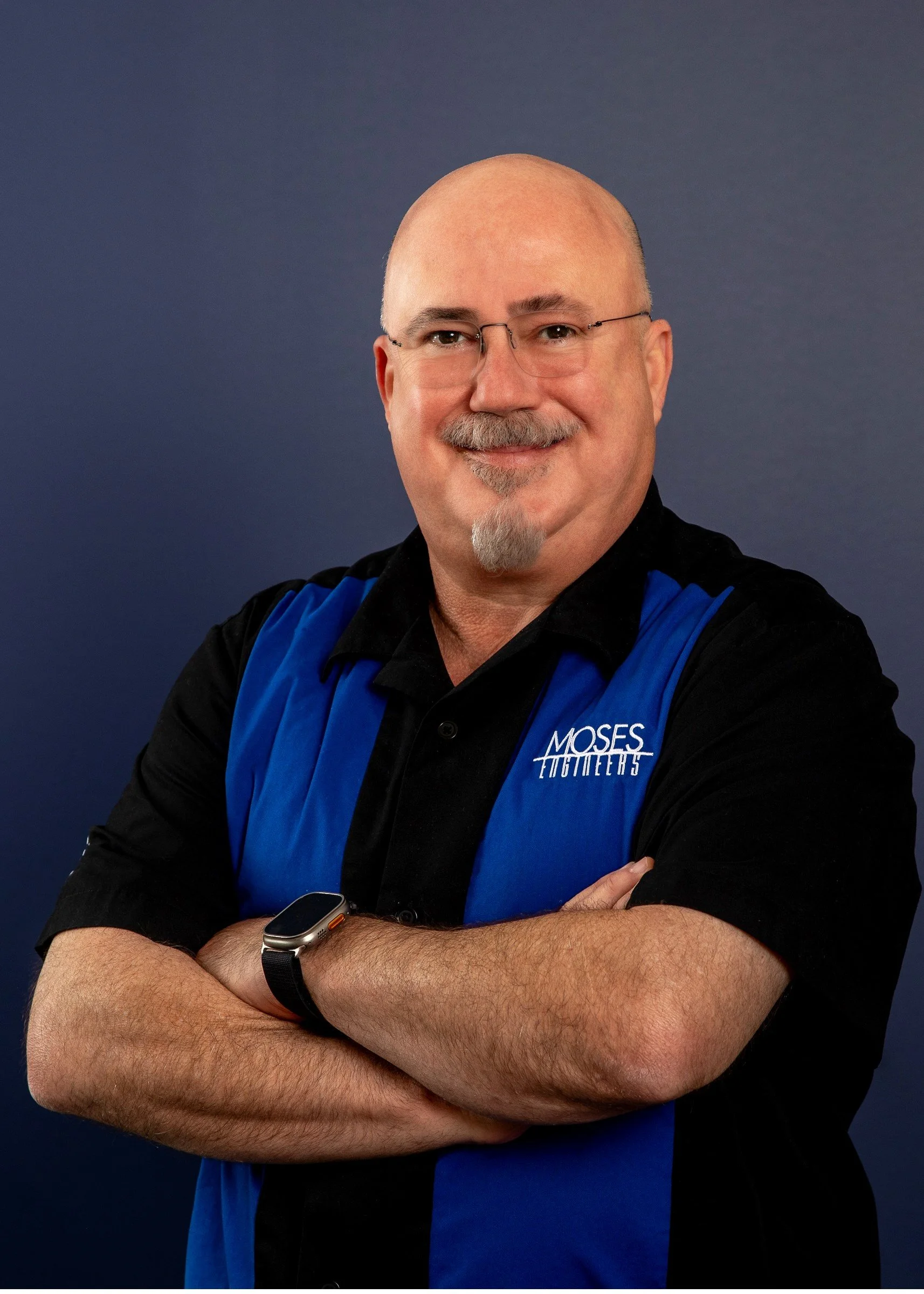 A middle-aged man with glasses, a bald head, and a mustache and goatee, smiling with arms crossed, wearing a black and blue shirt with "Mosess Engineers" embroidered on it, standing against a dark blue background.