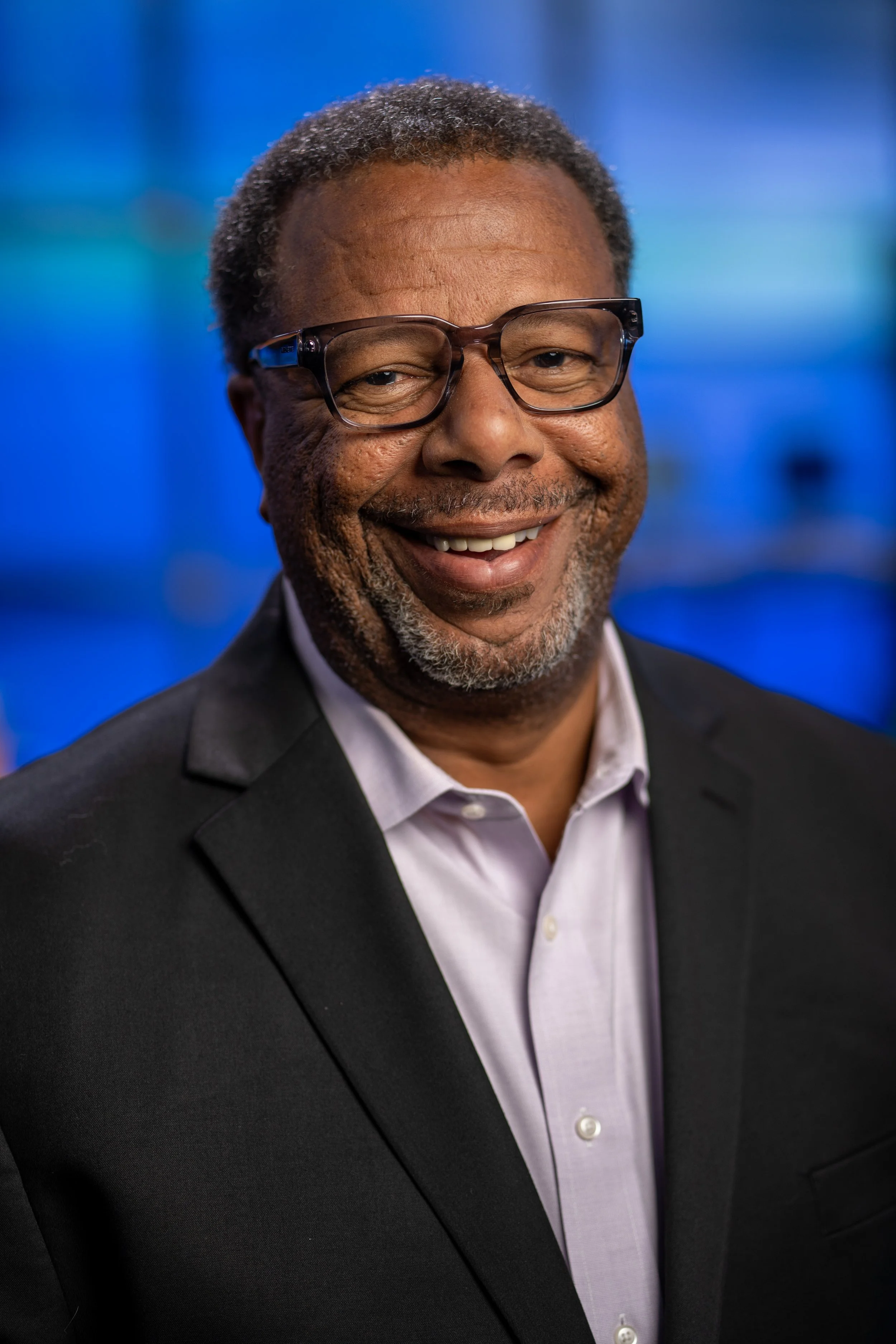 A smiling middle-aged African American man with gray hair and glasses, wearing a black suit and white shirt, against a blurred blue background.