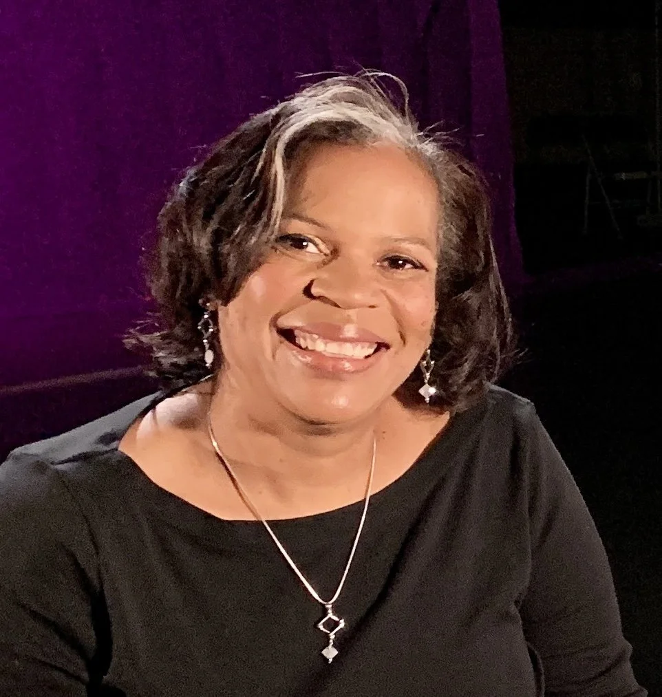 A smiling woman with short curly hair, wearing a black top and jewelry, sitting against a dark purple background.