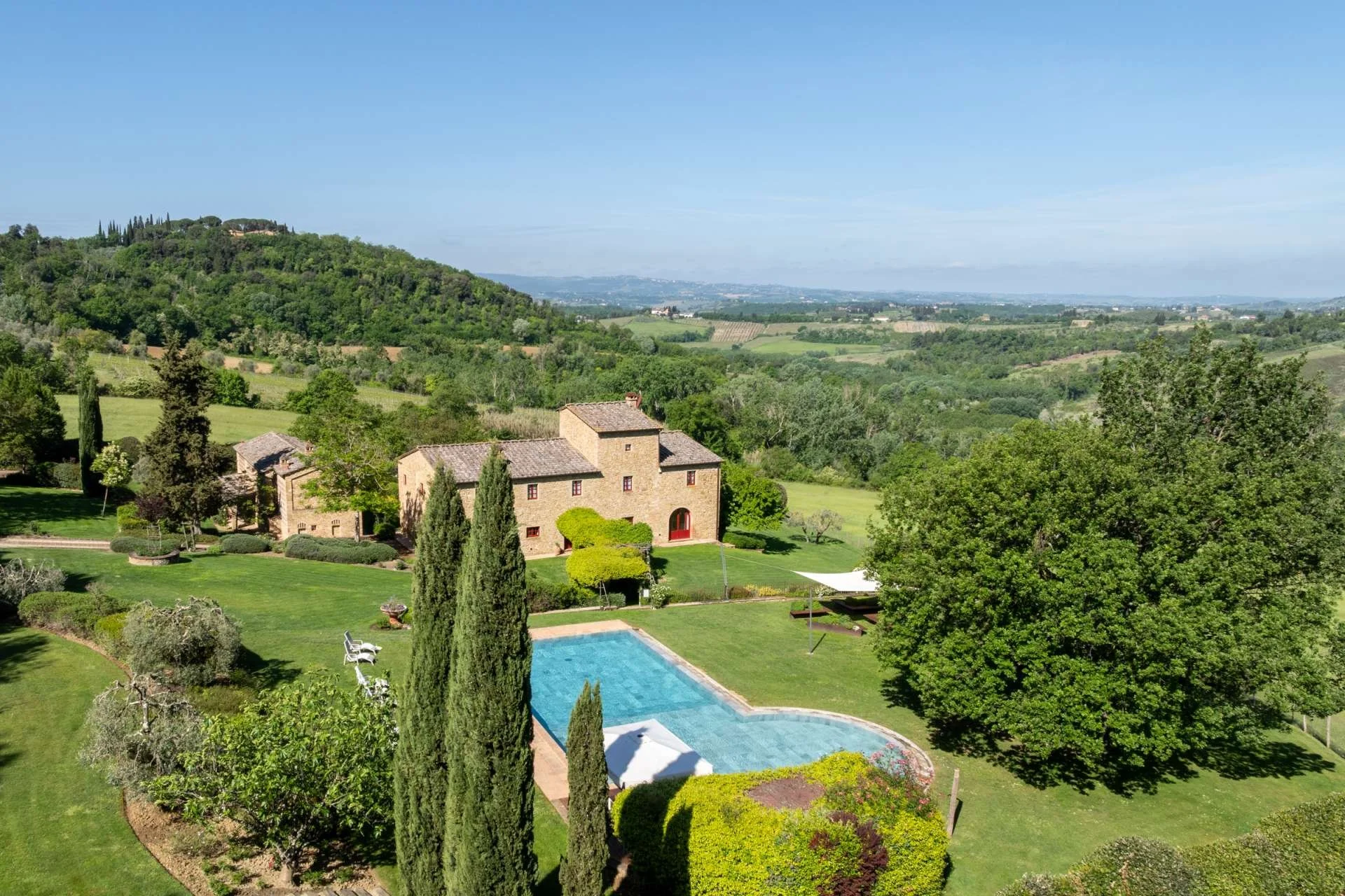 Landscape view of a lush, green estate with a stone house, swimming pool, surrounded by tall trees, rolling hills, and farmland in the distance under a clear blue sky.