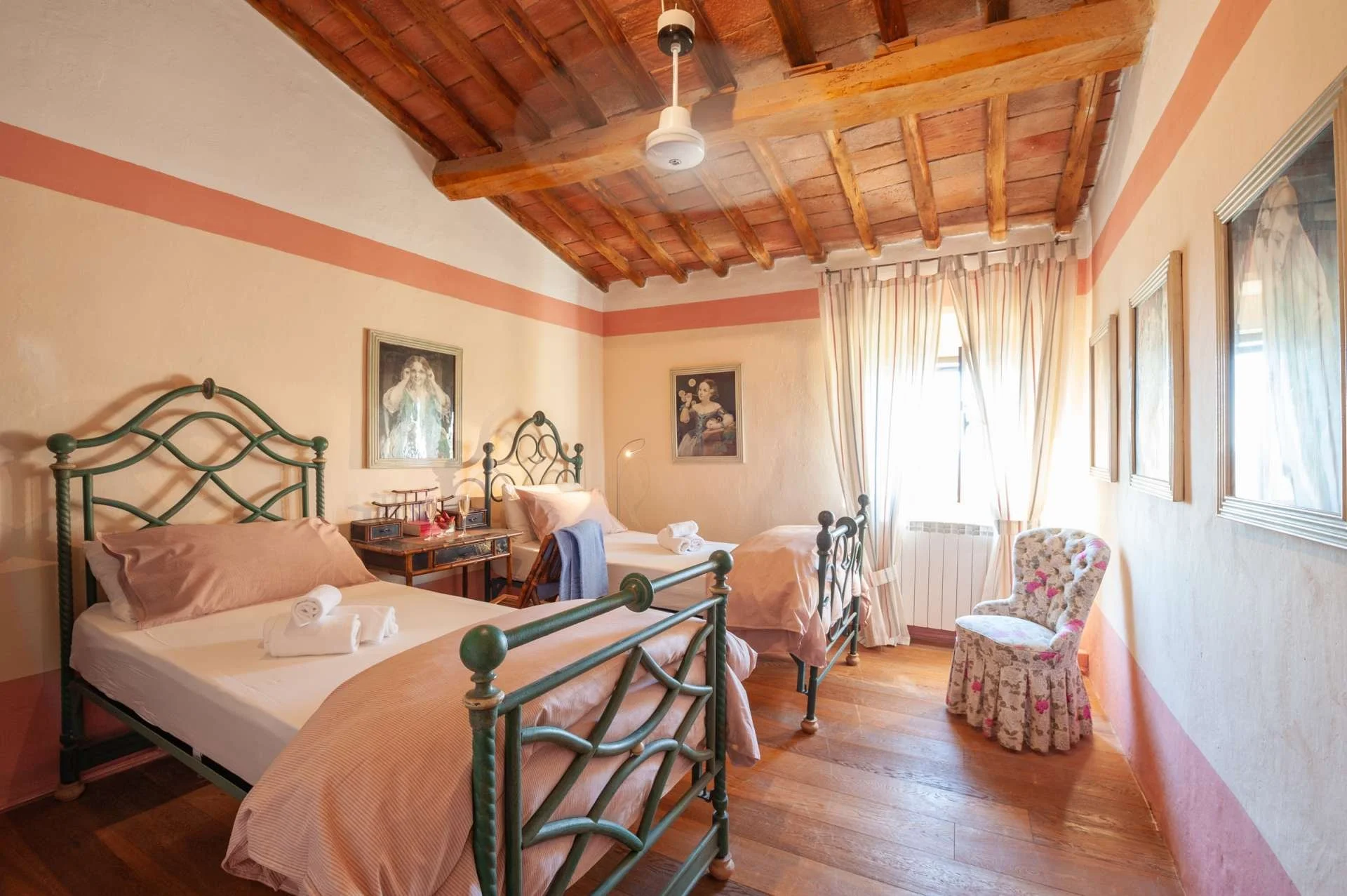 A cozy bedroom with three metal framed beds, each with beige bedding. Decor includes framed art on the light-colored walls, a wooden dressing table between two beds, and a floral armchair by a window with floral curtains. A ceiling fan with a light fixture is mounted on a wooden ceiling with exposed beams.