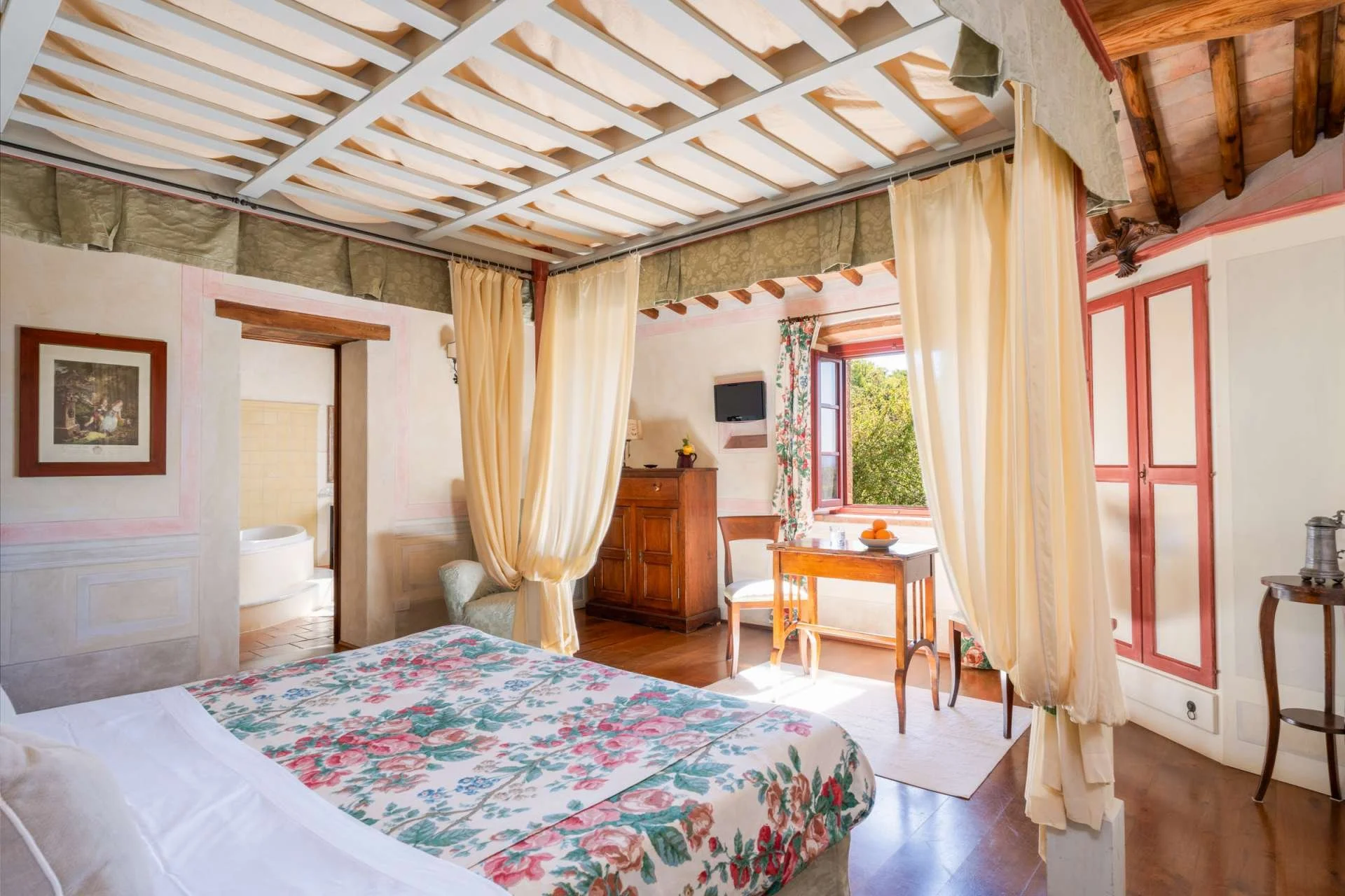 Interior of a cozy bedroom with a floral bedspread, cream-colored curtains, wooden furniture, and an open window showing lush greenery outside.